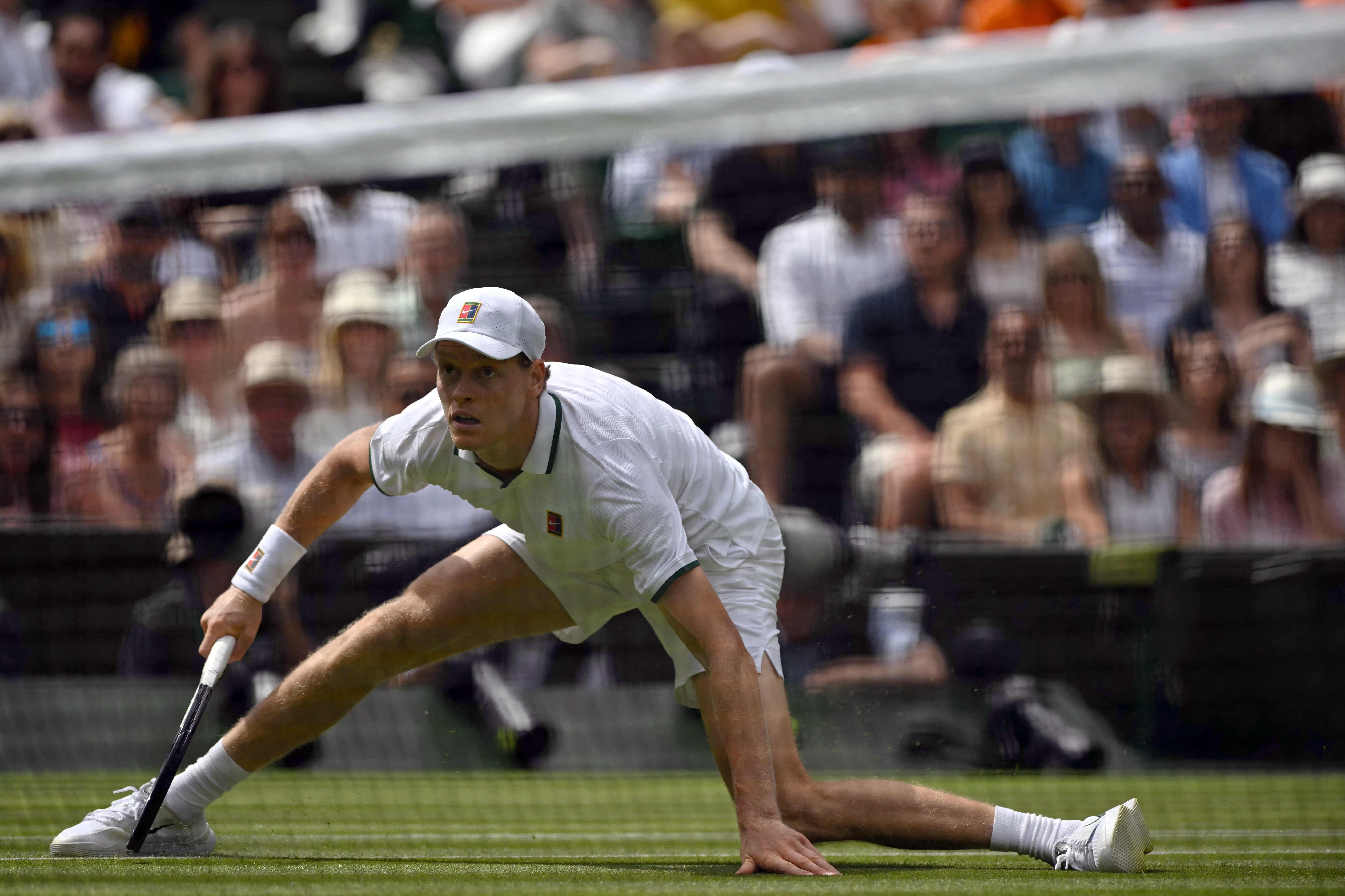 Tennisspieler in weißer Kleidung streckt sich am Boden, um einen Ball auf Rasen zu erreichen.