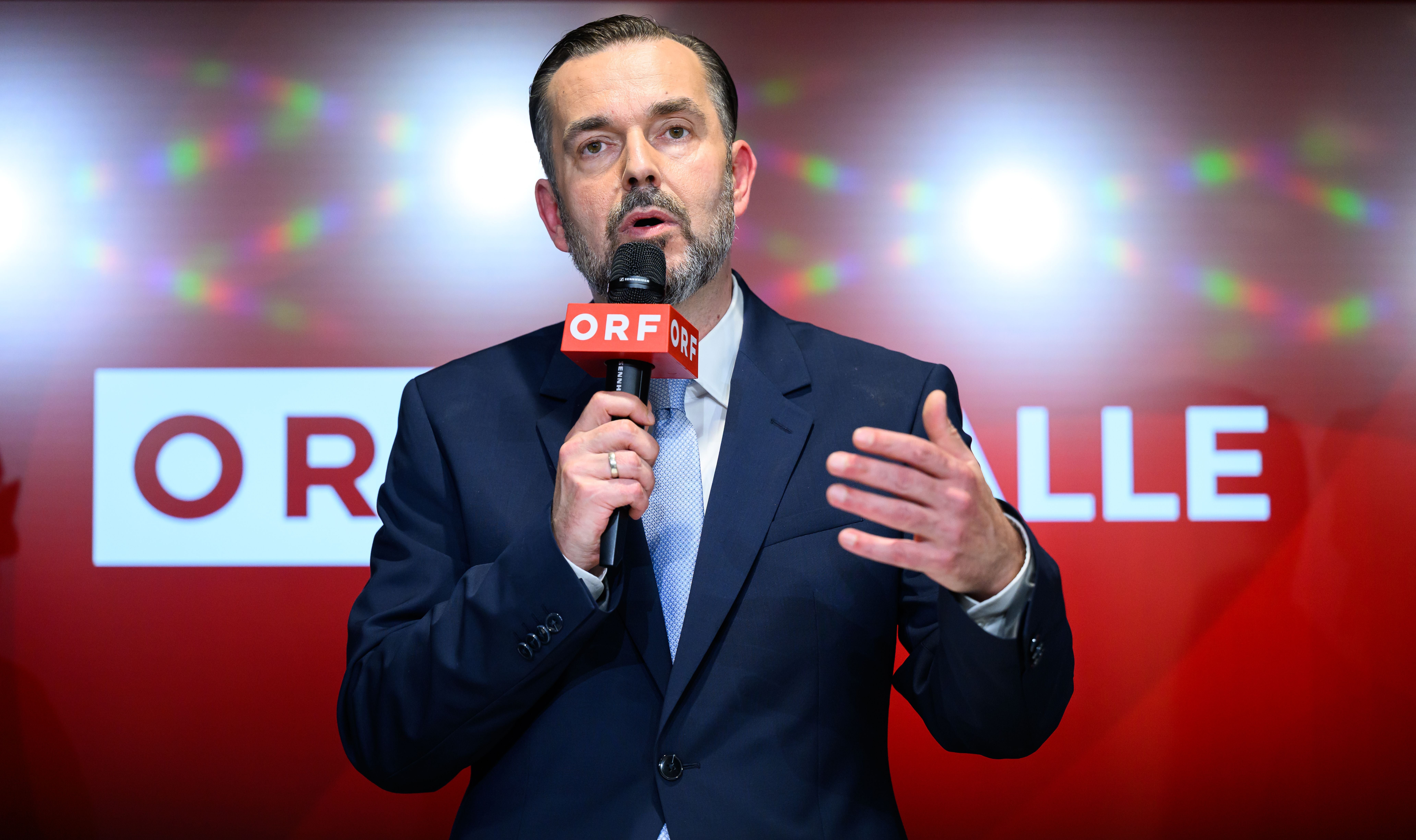 Ein Mann im Anzug spricht mit ORF-Mikrofon vor rotem Hintergrund bei einer Veranstaltung.