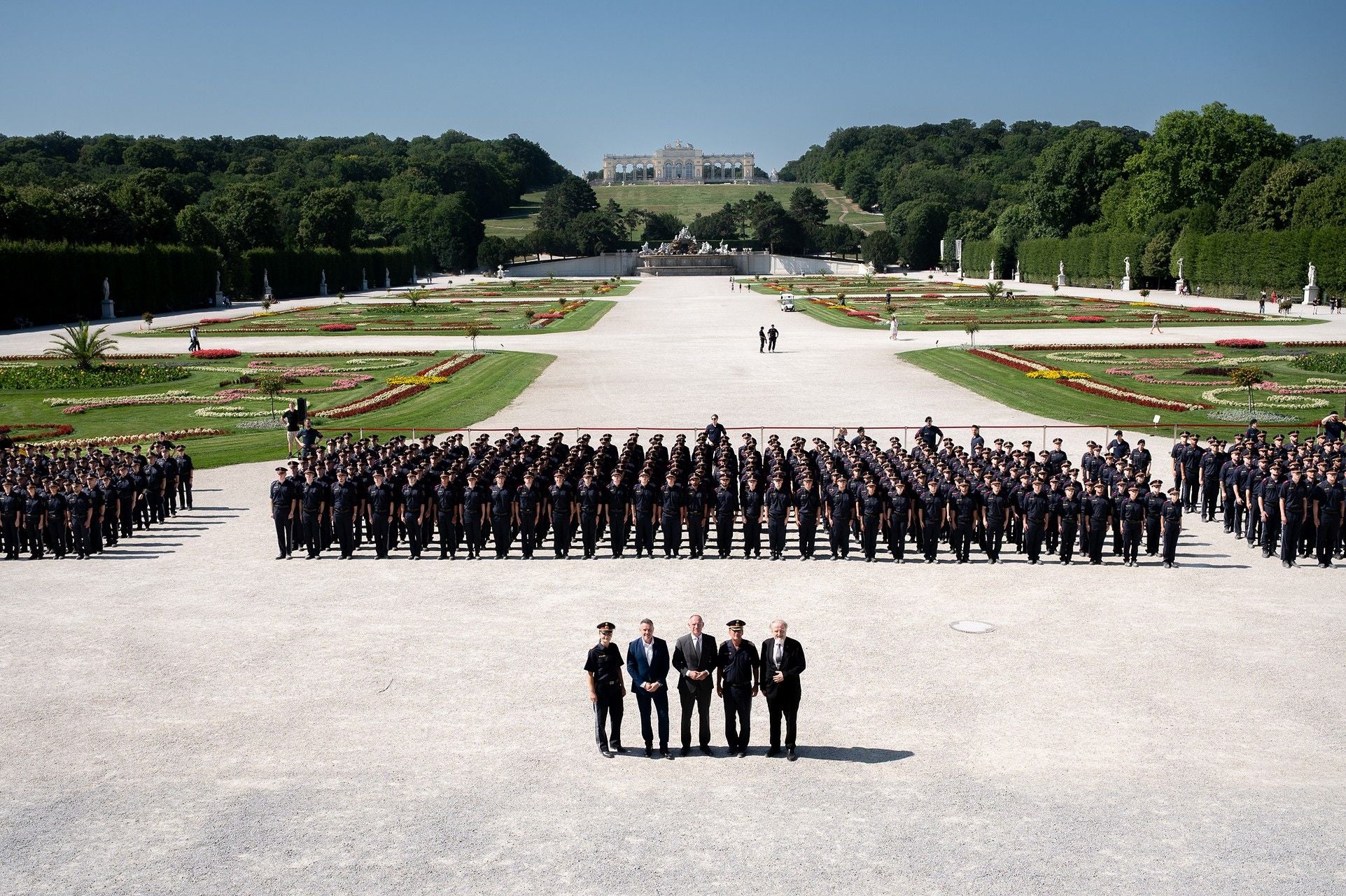 Große Gruppe uniformierter Personen steht in Reihen in einem Schlossgarten, vorne fünf Männer.