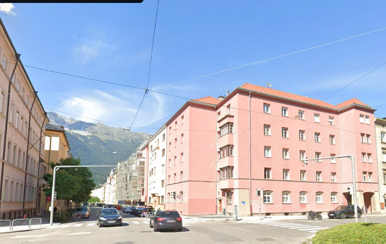 Kreuzung mit Autos und rosa Wohngebäude in der Speckbacher Straße in Innsbruck bei Sonnenschein.