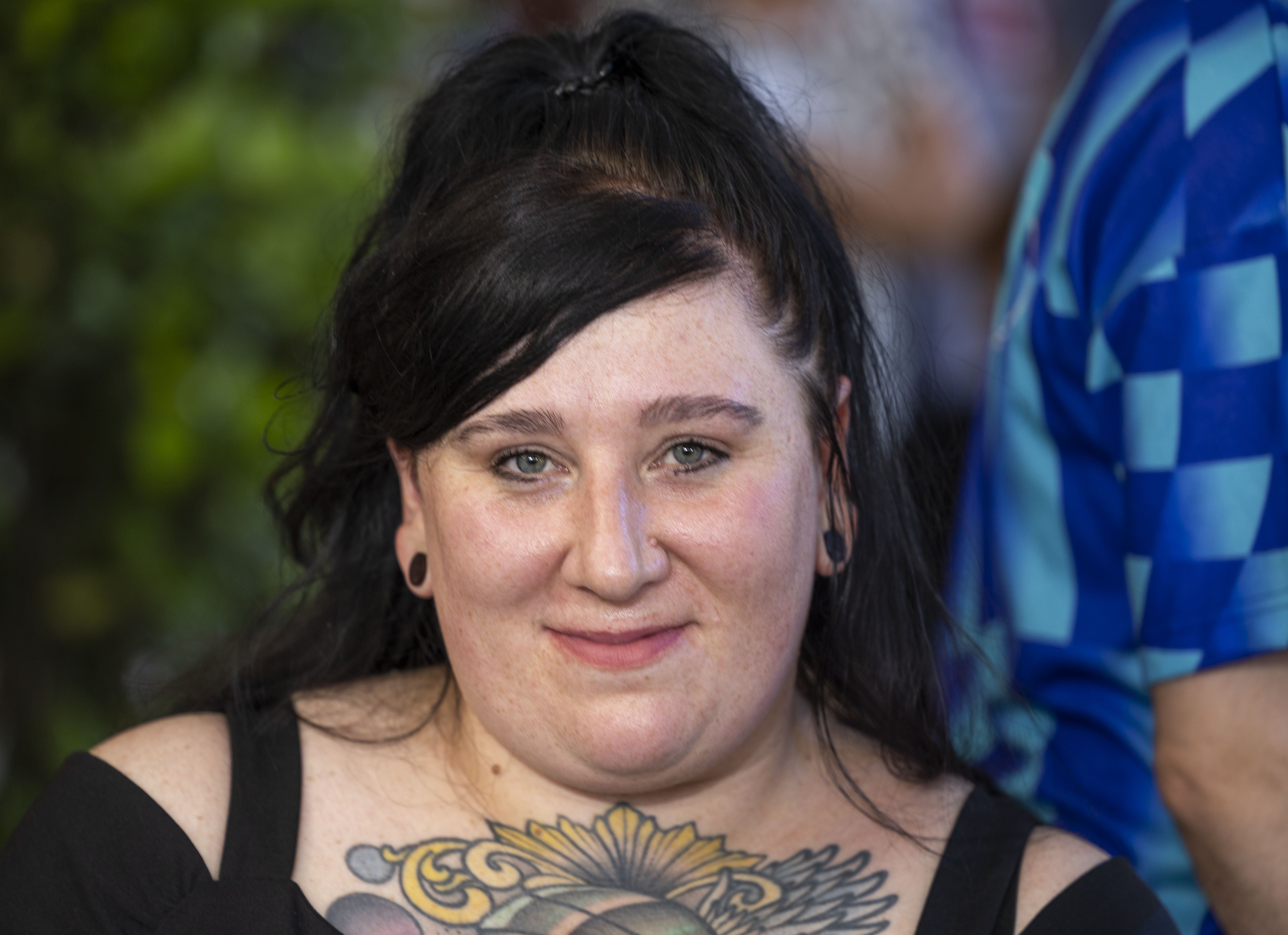 Junge Frau mit schwarzen Haaren und großem Brust-Tattoo lächelt in die Kamera.