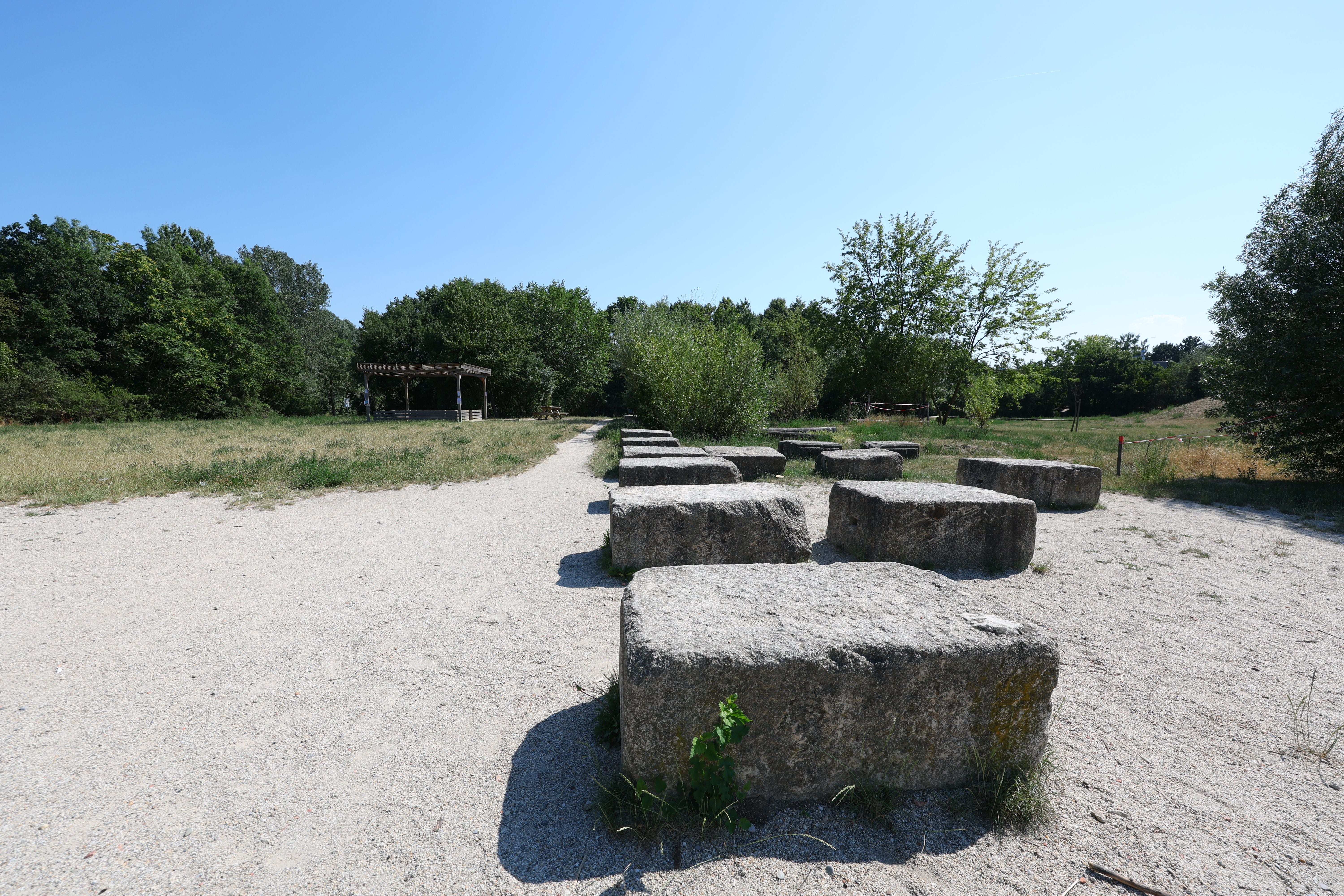 Große Steinquader stehen auf einer sandigen Fläche im Park, umgeben von Bäumen und einem Pavillon.