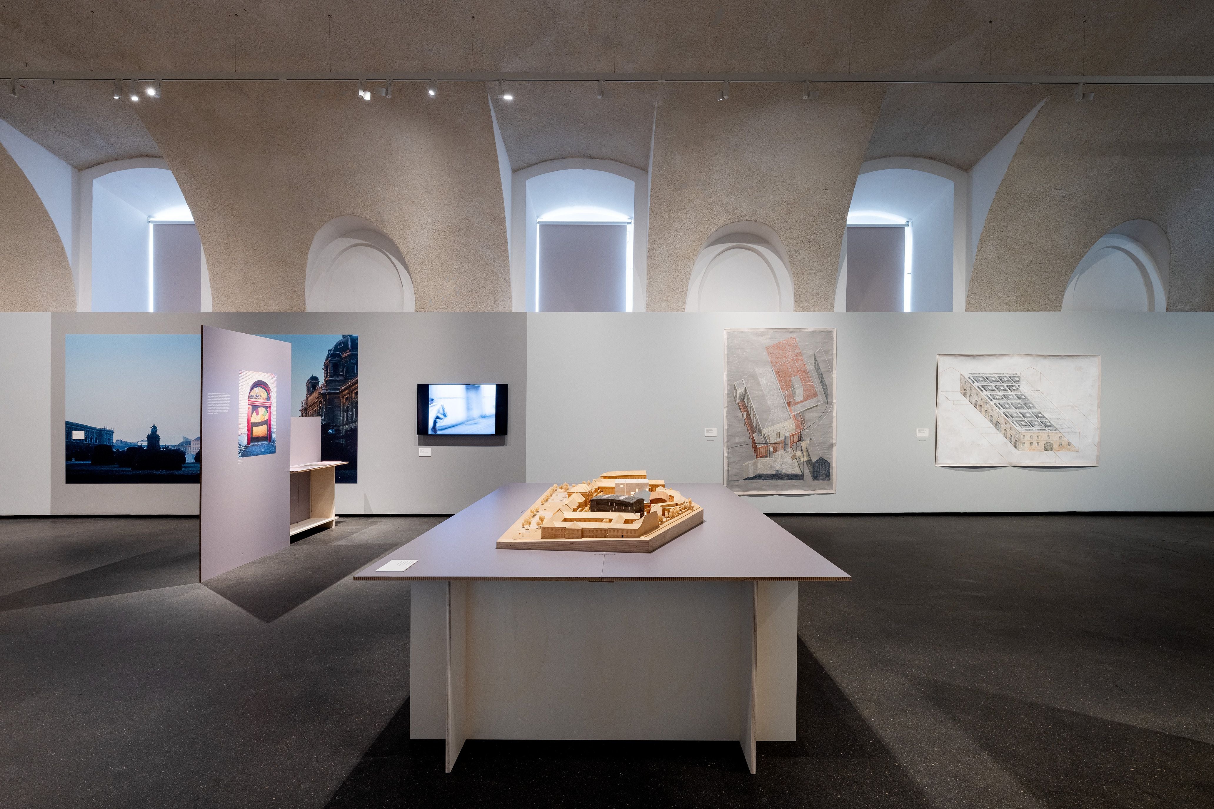 Ausstellung mit Architekturmodell auf Tisch, Zeichnungen und Fotos an neutralgrauen Wänden.