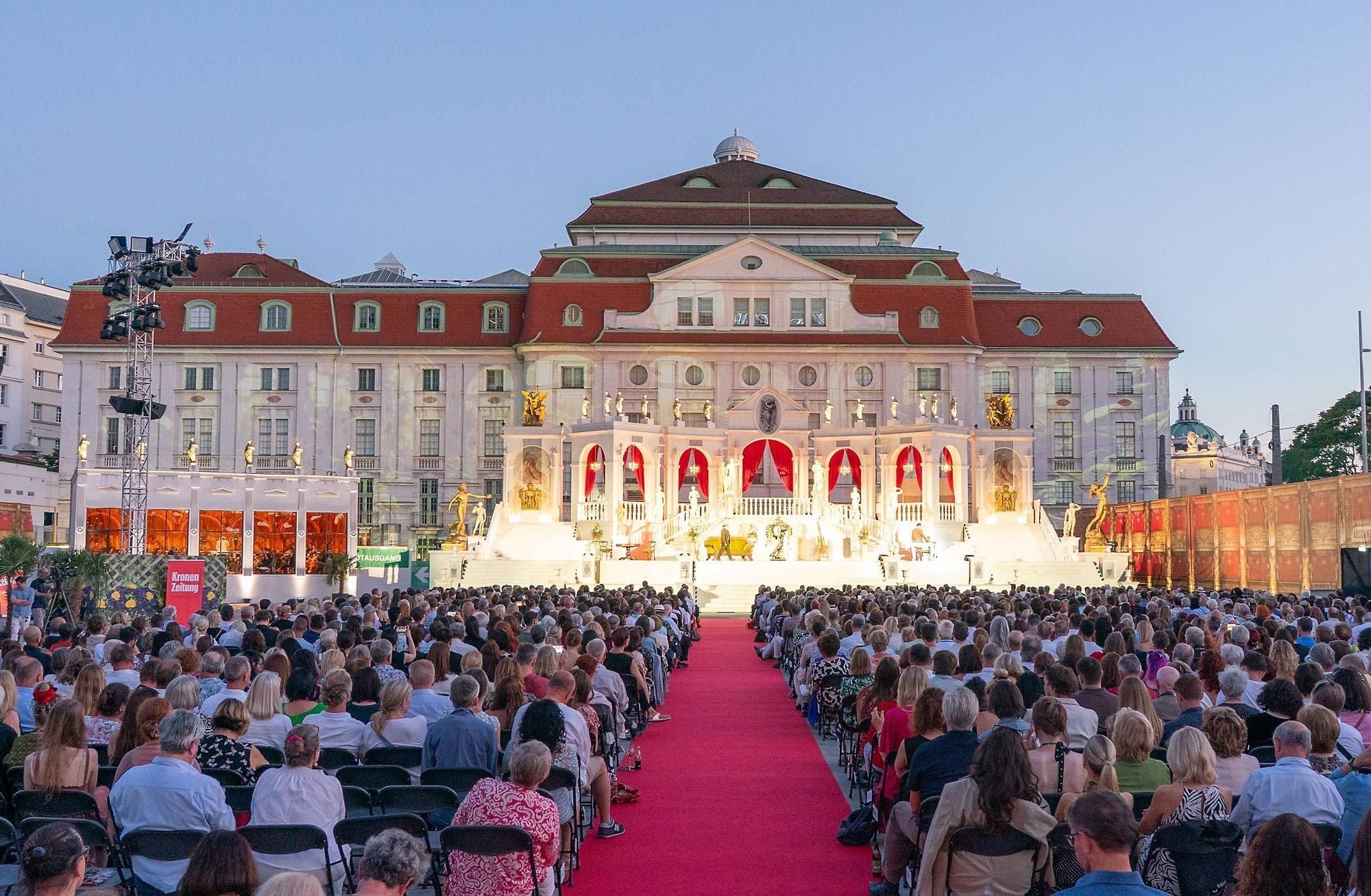 Open-Air-Opernaufführung mit großem Publikum vor einem festlich beleuchteten Gebäude.