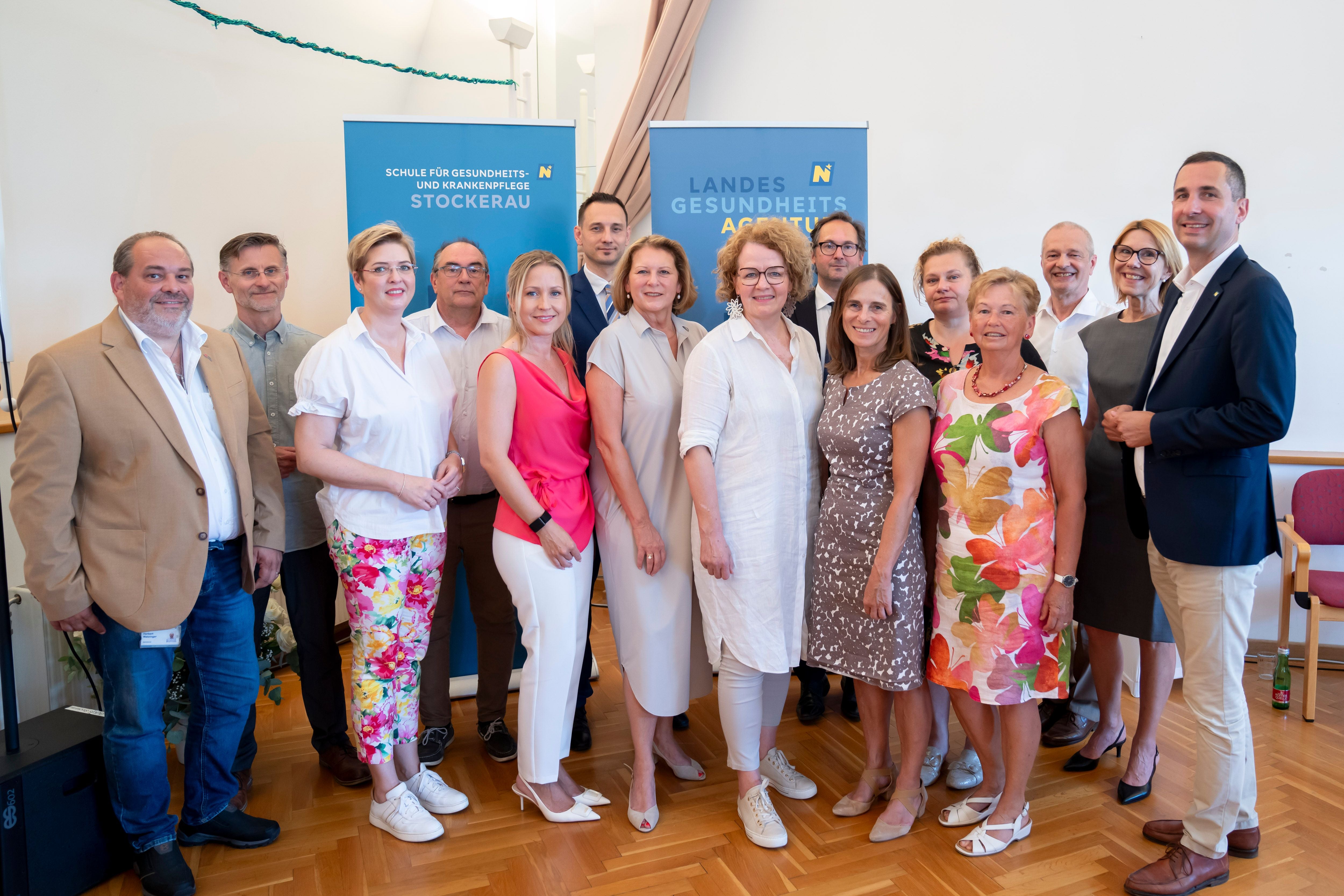 Gruppenfoto von 16 Personen vor zwei blauen Bannern in einem hellen Raum mit Holzfußboden.