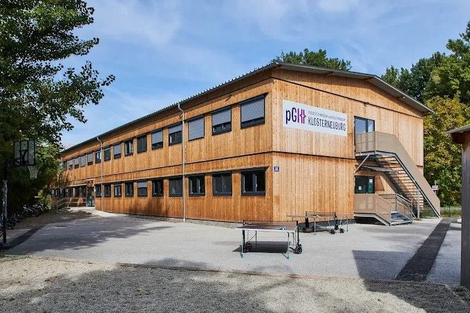 Zweistöckiges Schulgebäude aus Holz mit Außentreppe und Tischtennisplatte davor.