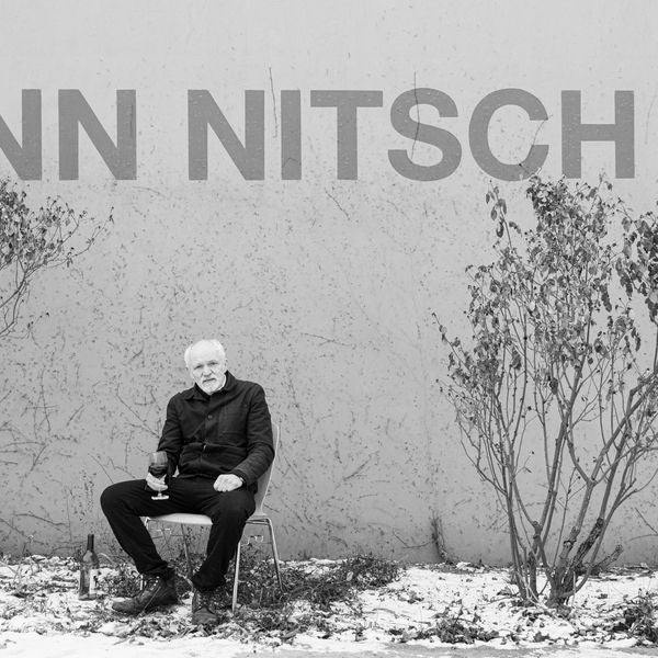 Ein älterer Mann sitzt mit Weinglas vor einer Wand mit Aufschrift „NITSCH“, winterliche Umgebung.