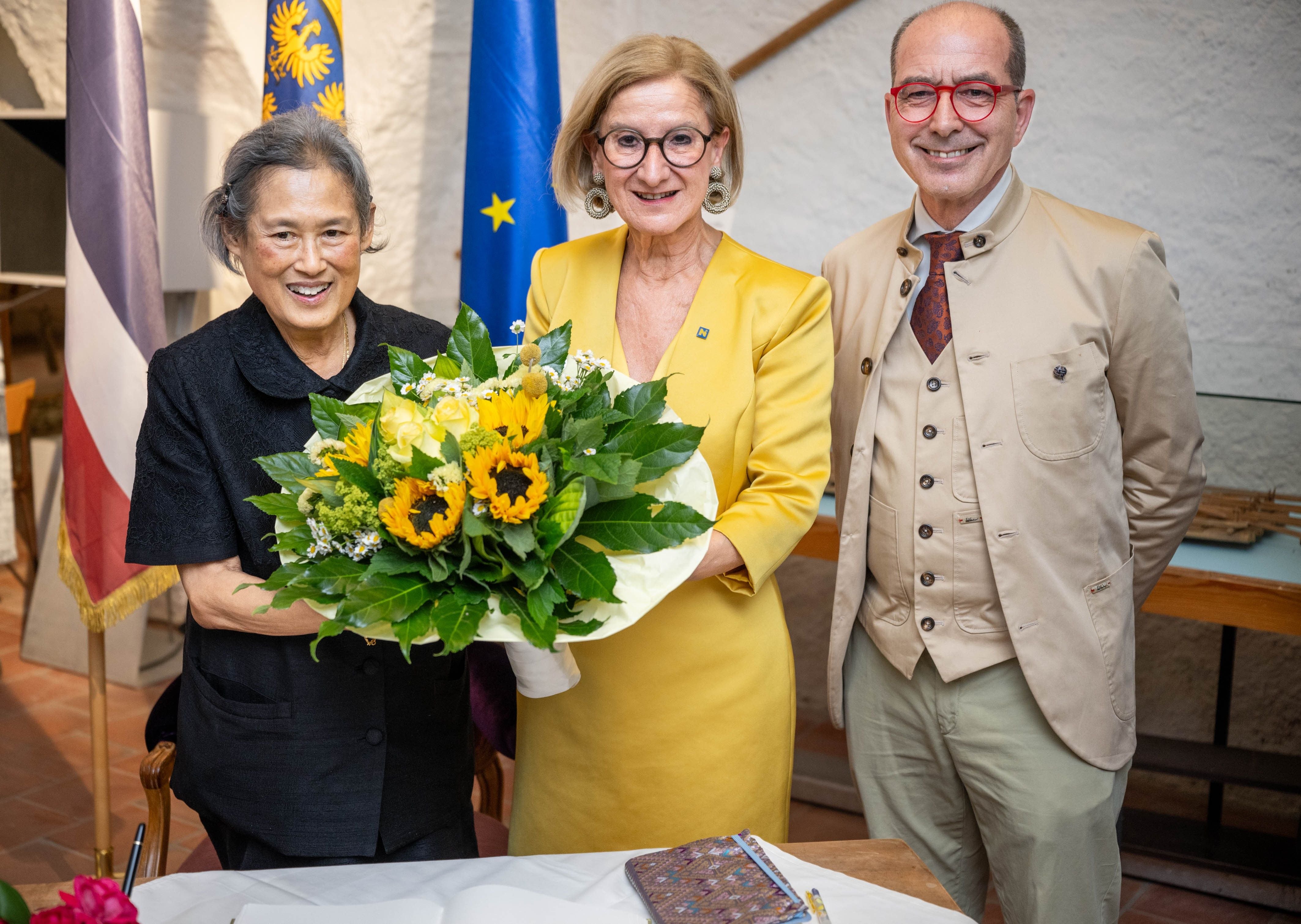 Drei Personen posieren mit einem Strauß Sonnenblumen vor Flaggen und einem Tisch.