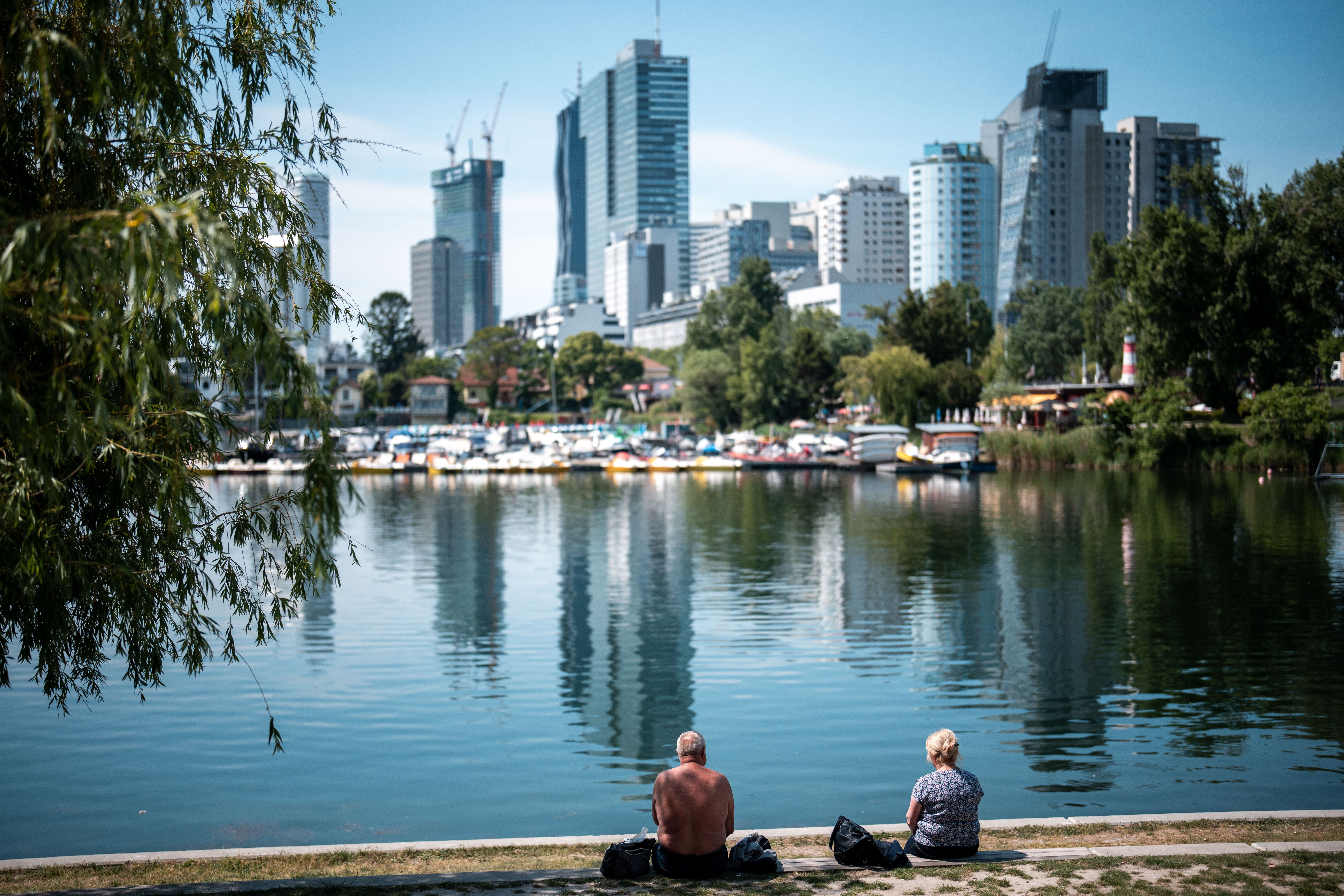 Zwei Personen sitzen am Ufer der Alten Donau, im Hintergrund Wiens moderne Skyline.