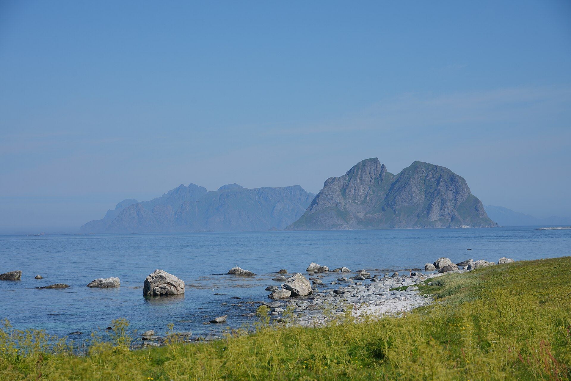Felsige Küste mit Blick auf die Insel Mosken im ruhigen blauen Meer, im Hintergrund Berge.