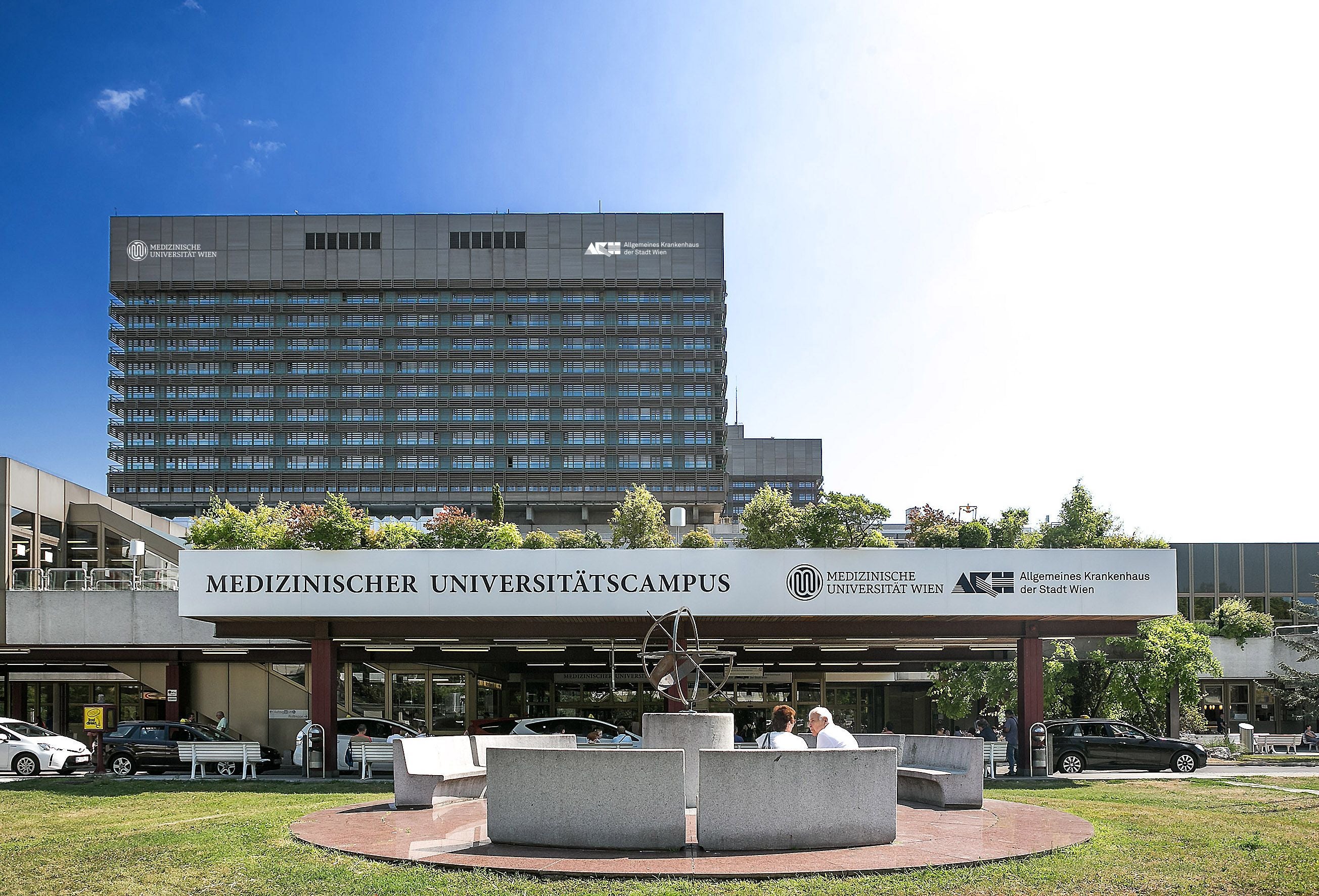 Eingangsbereich des MedUni Wien AKH mit moderner Fassade und Menschen auf Bänken im Vordergrund.