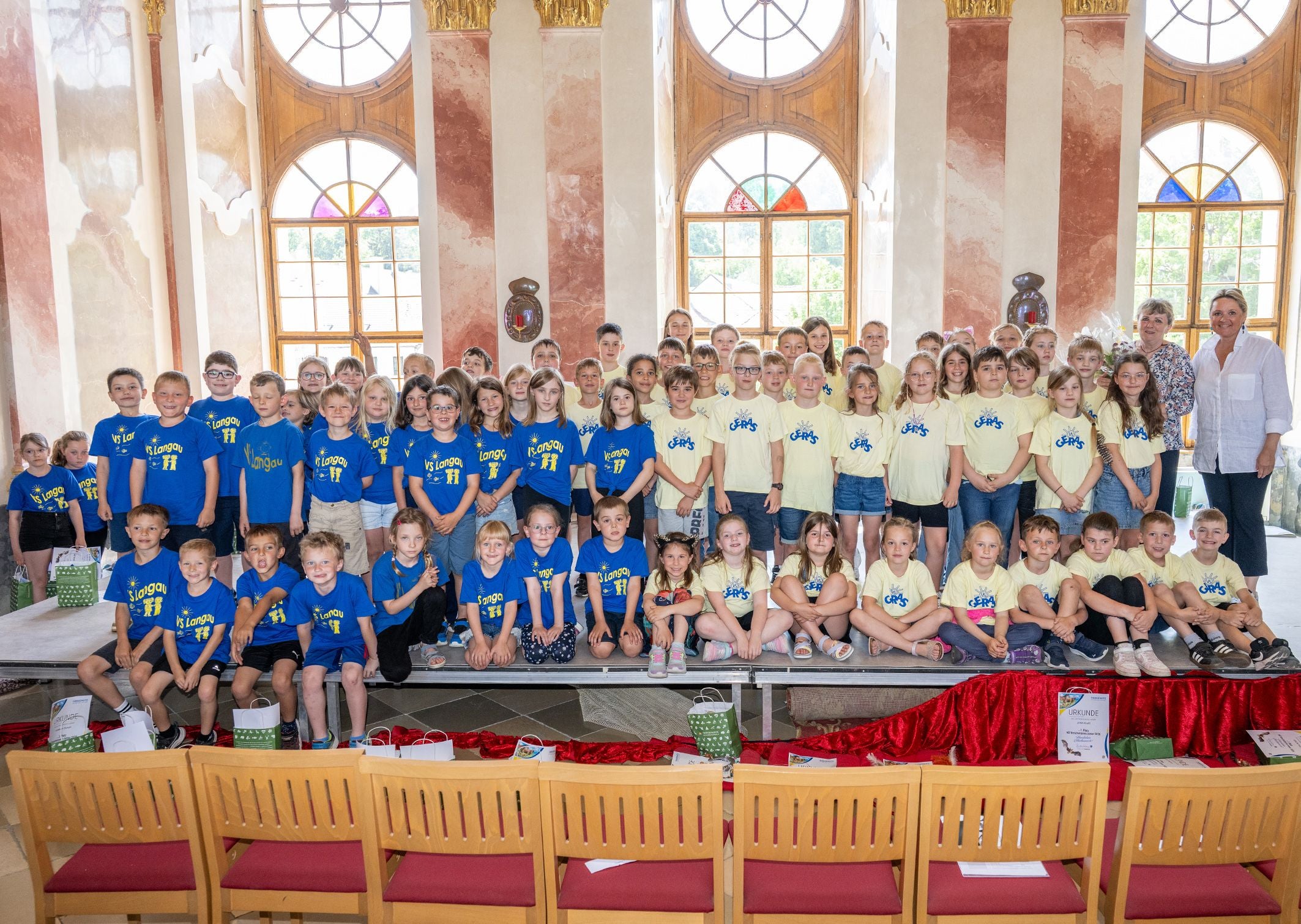 Gruppenfoto vieler Kinder in gelben und blauen T-Shirts in einem hellen, festlich dekorierten Saal.