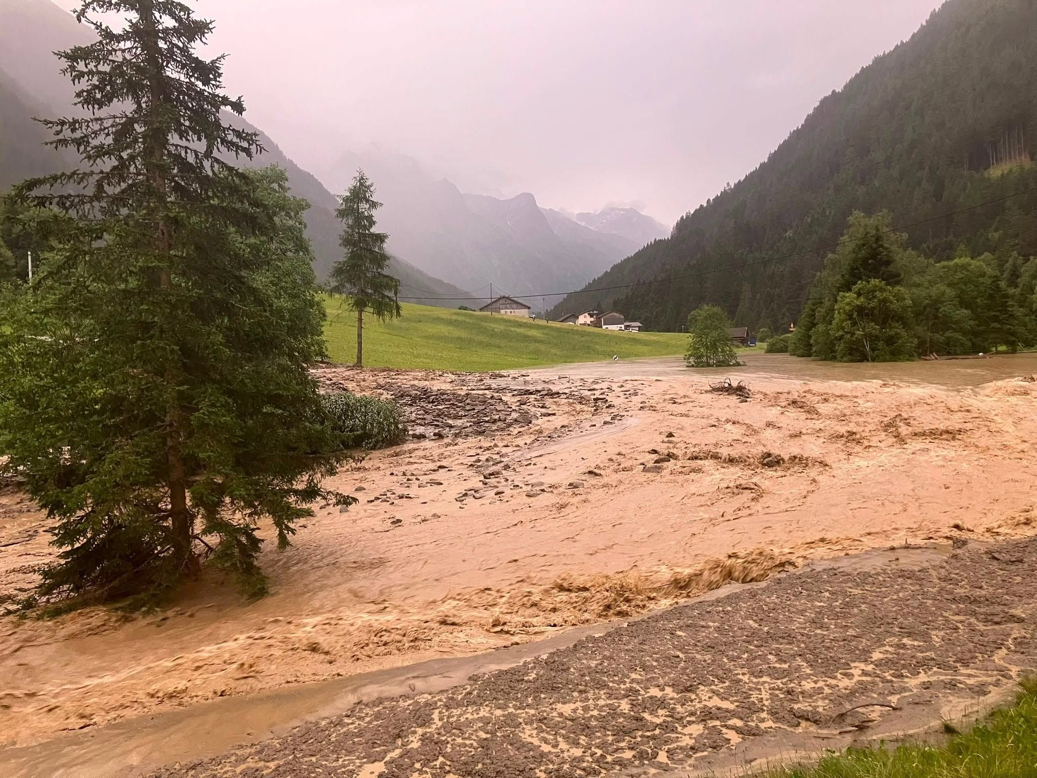 Starkes Hochwasser überflutet eine Wiese bei Bäumen im Tal, Berge im Hintergrund.