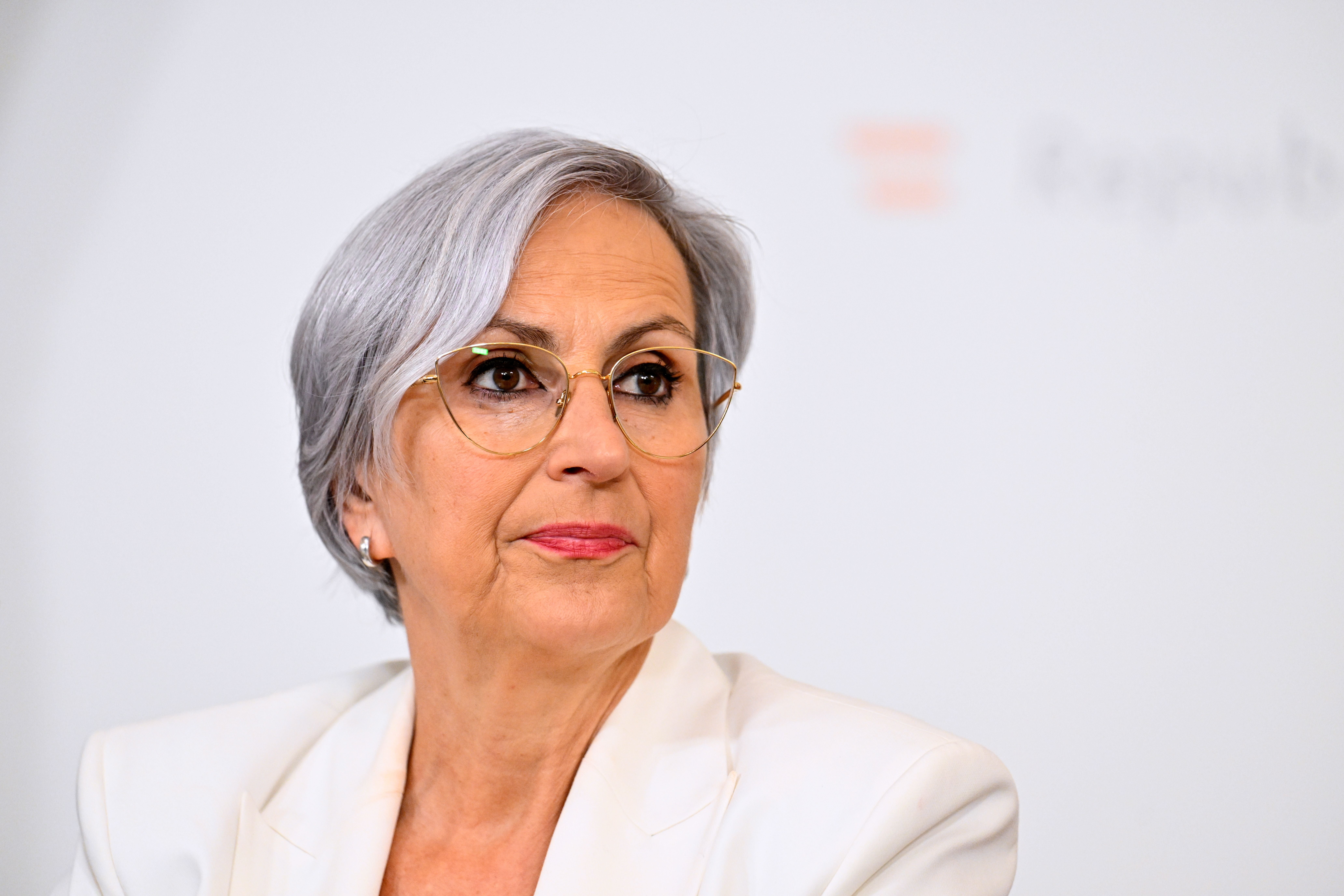 Eine ältere Frau mit kurzen grauen Haaren und Brille trägt einen weißen Blazer.