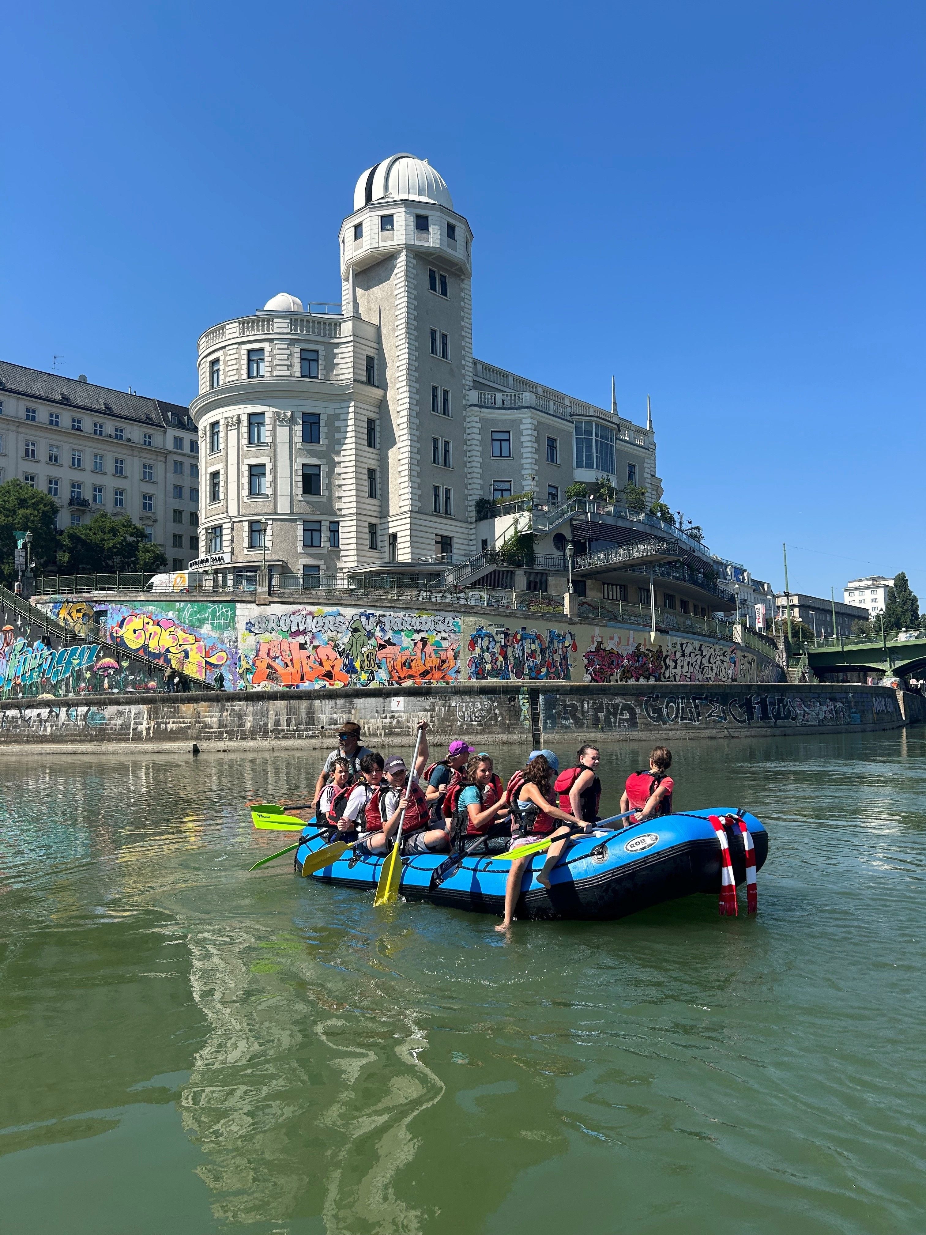 Eine Gruppe Menschen paddelt in einem Schlauchboot auf dem Donaukanal vor graffiti-bedeckten Wänden.