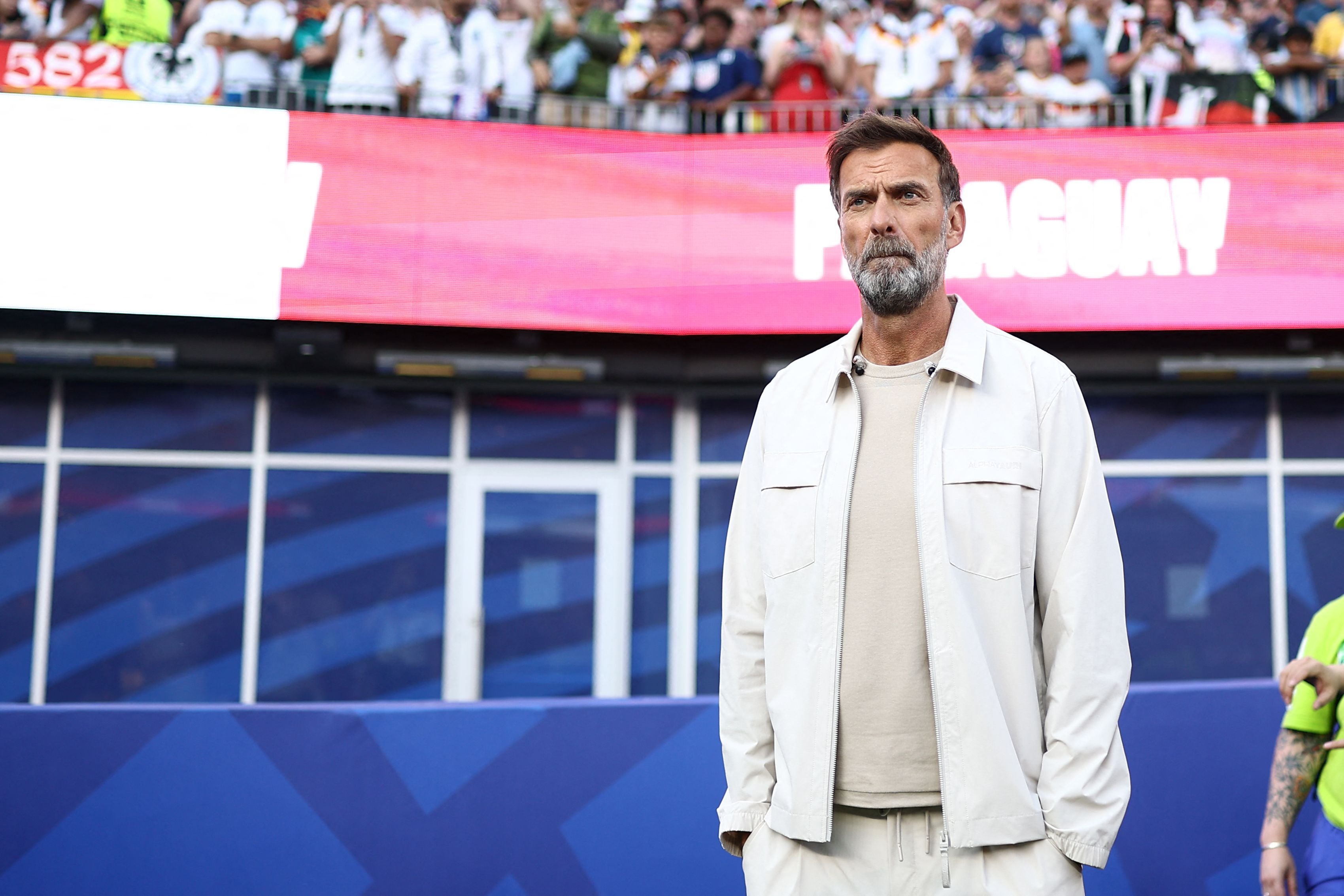 Ein Mann im hellen Outfit steht nachdenklich in einem Fußballstadion vor einer Menschenmenge.