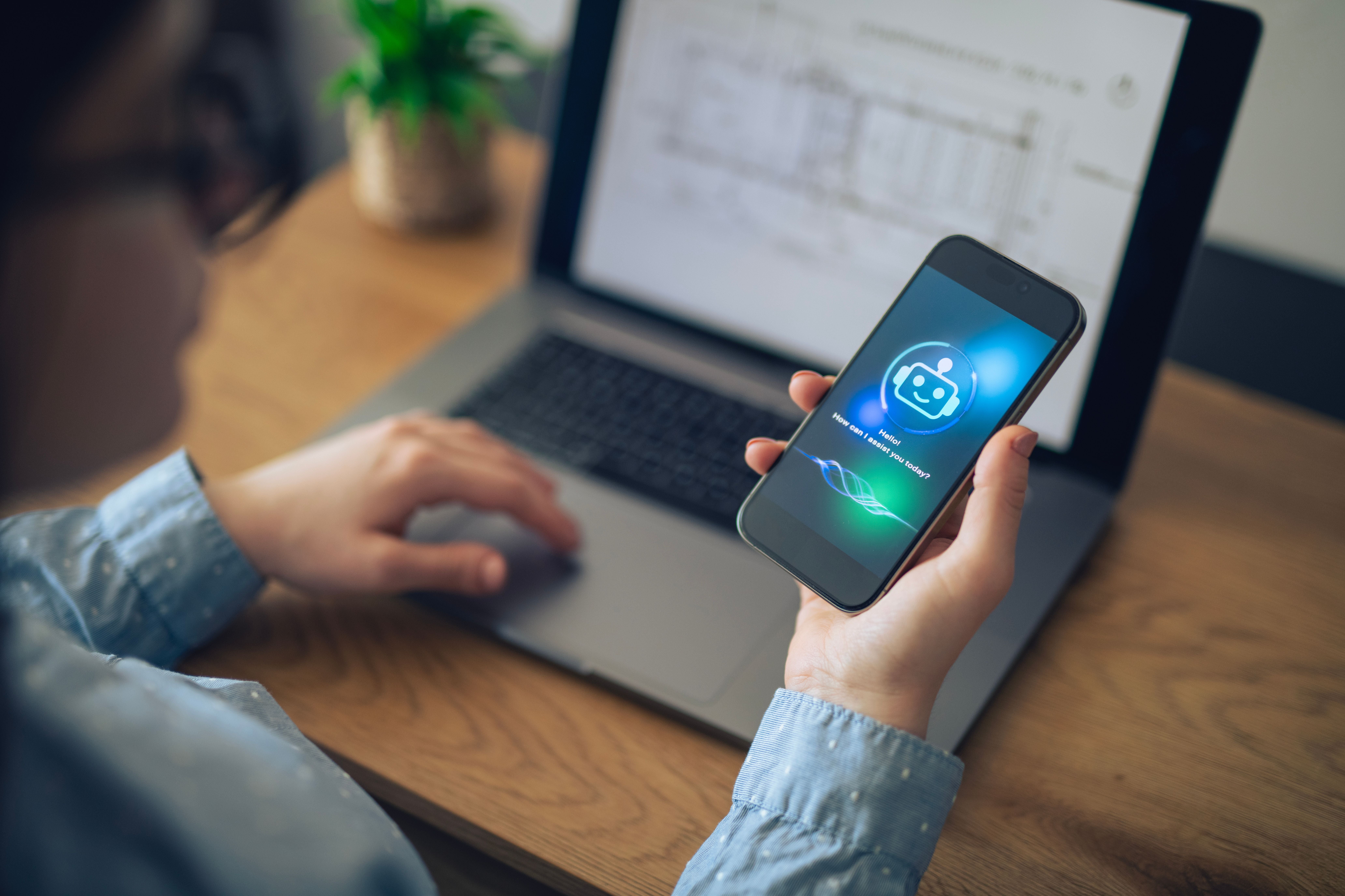 Frau nutzt einen KI-Chatbot auf ihrem Smartphone vor einem Laptop am Schreibtisch.