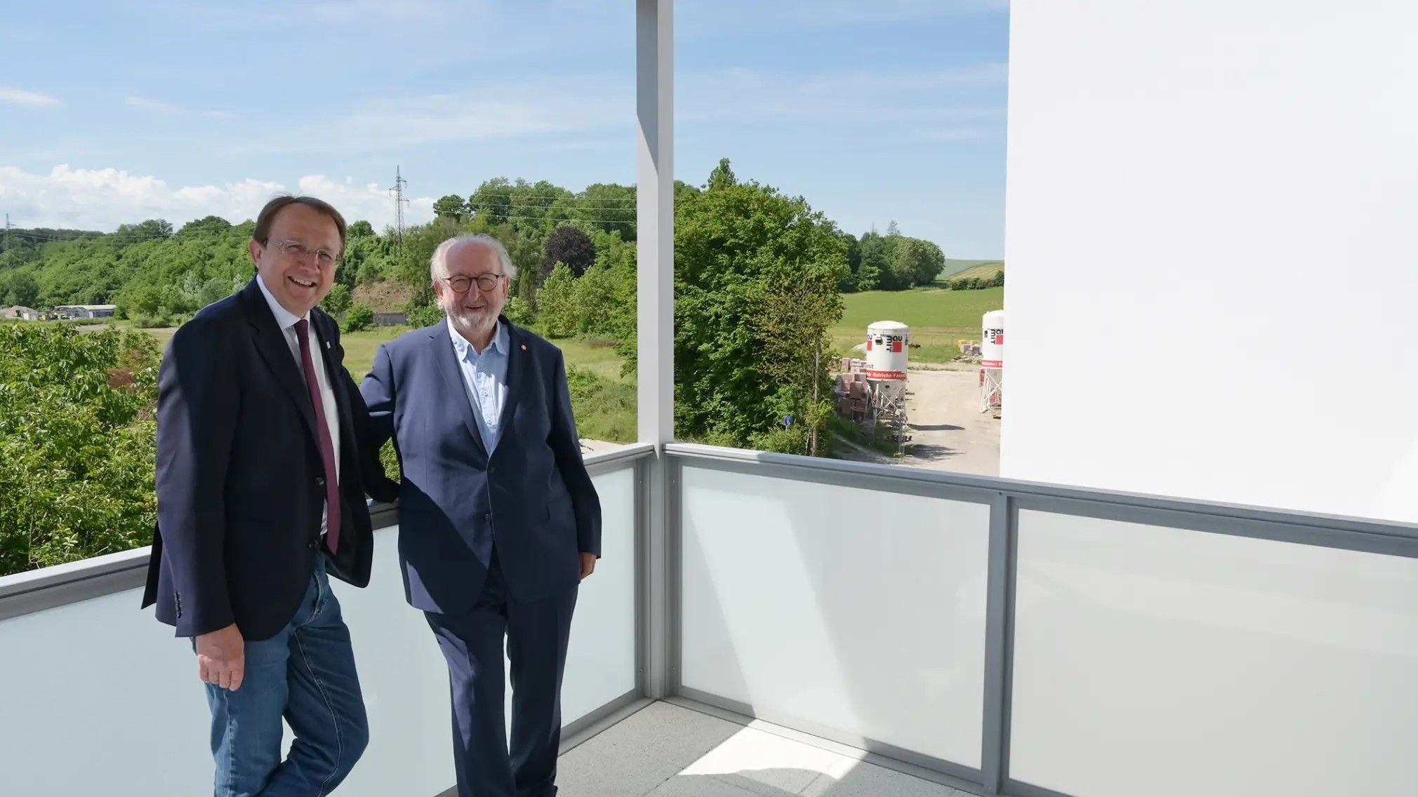 Zwei Männer im Anzug stehen auf einem Balkon und blicken auf eine Baustelle im Grünen.