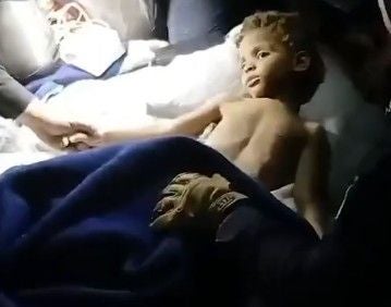 Ein kleiner Junge liegt gerettet und erschöpft unter einer Decke, umgeben von helfenden Händen.