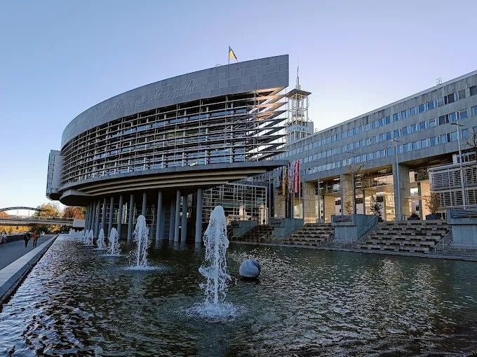 Modernes Parlamentsgebäude mit Springbrunnen davor und Landesflagge auf dem Dach.