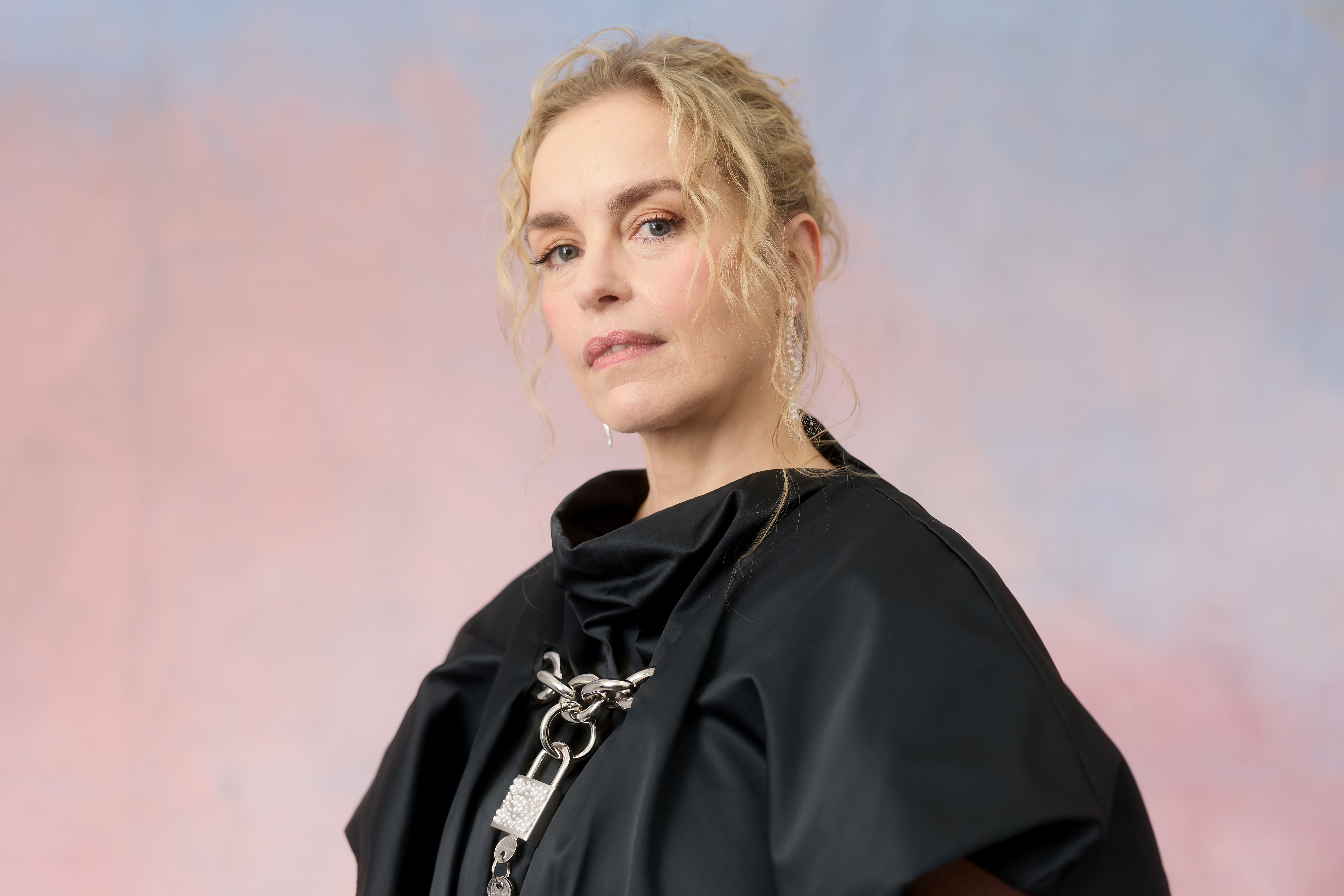 Frau mit blondem lockigem Haar und schwarzem Outfit vor pastellfarbenem Hintergrund.