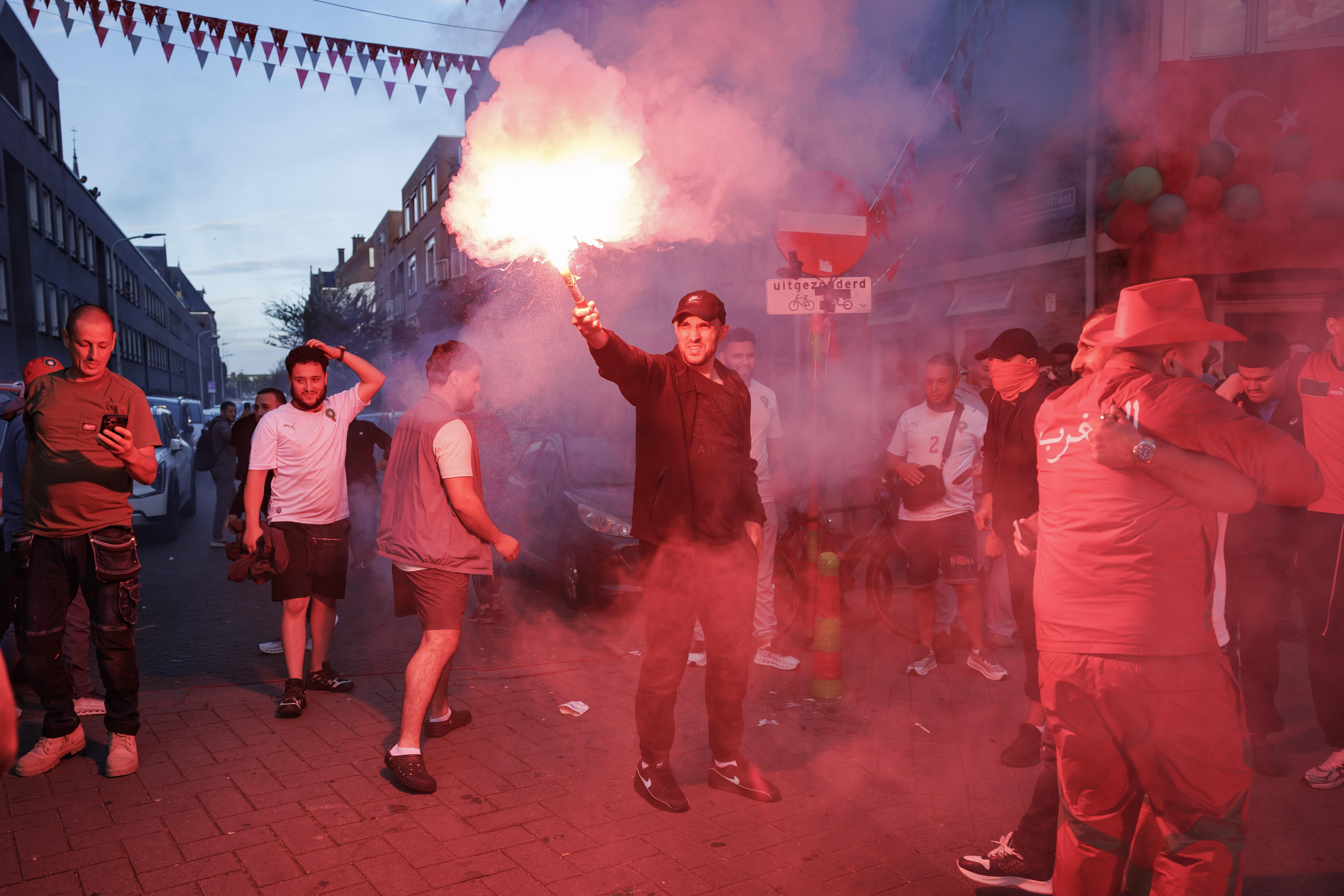 Fans feiern auf einer Straße, einer hält eine rote Rauchfackel, viele tragen Sportkleidung.