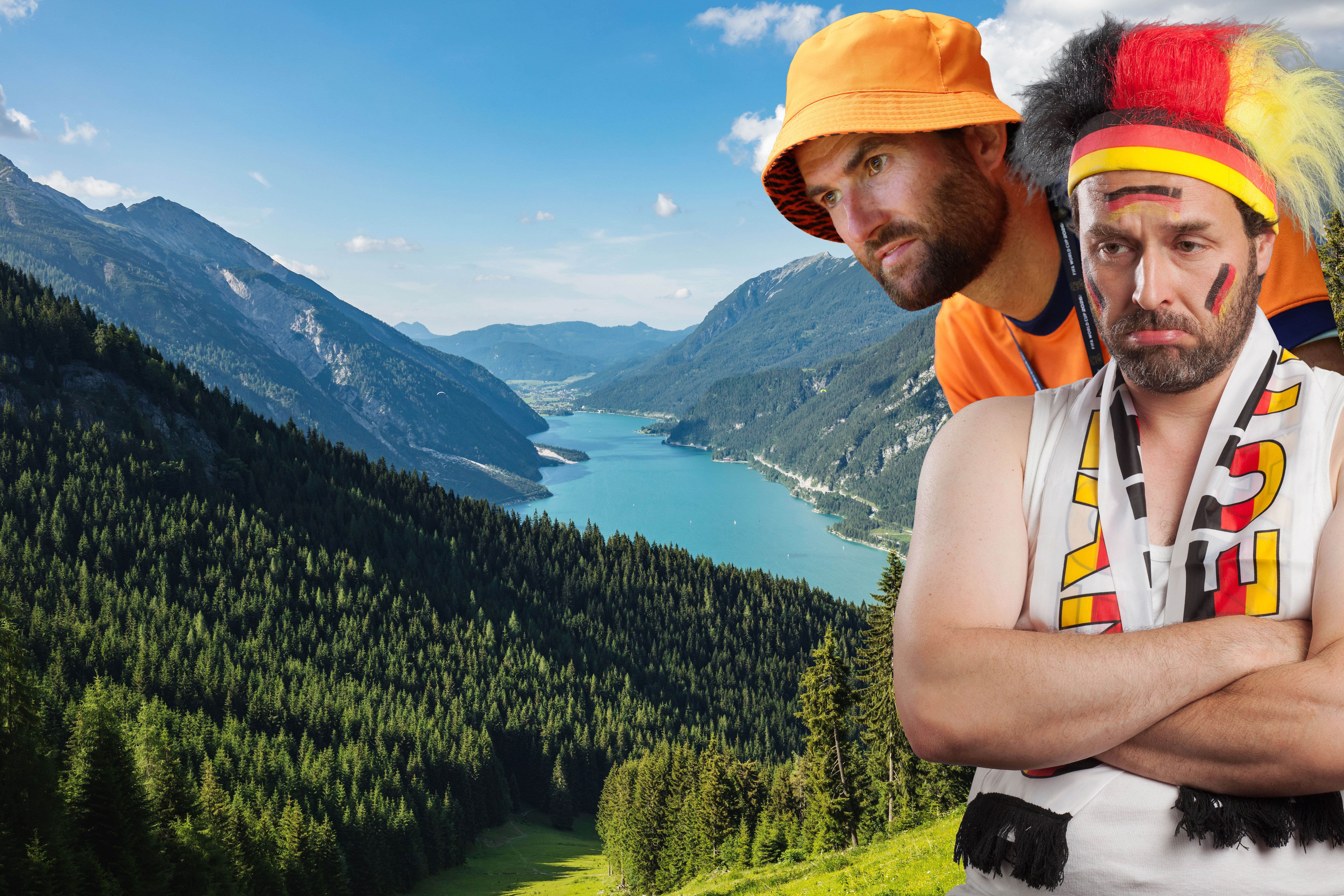 Zwei Fußballfans, einer für die Niederlande, einer für Deutschland, vor einer Alpenlandschaft.