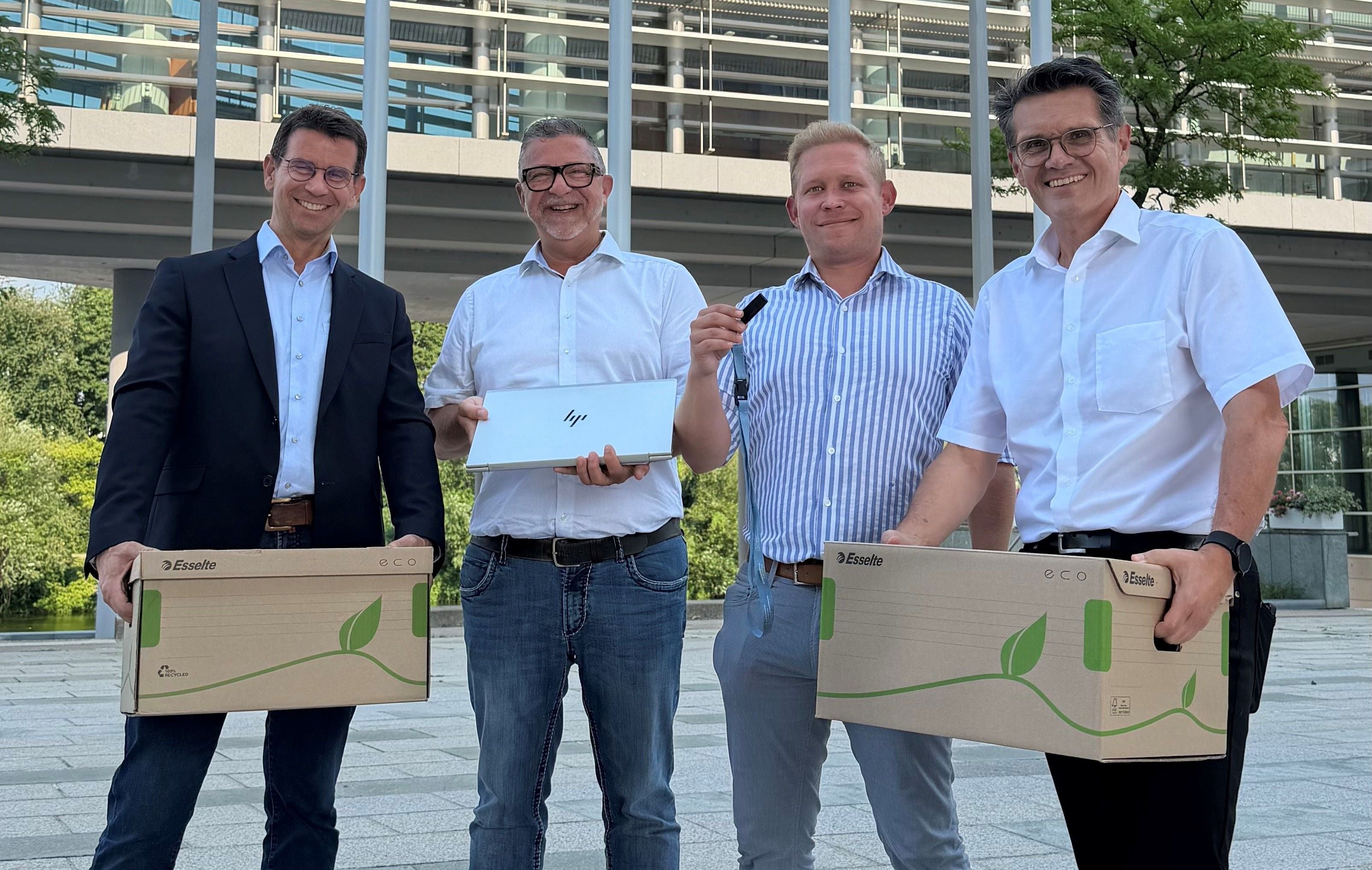 Vier Männer stehen draußen, halten Kartons, einen Laptop und einen USB-Stick, und lächeln in die Kamera.