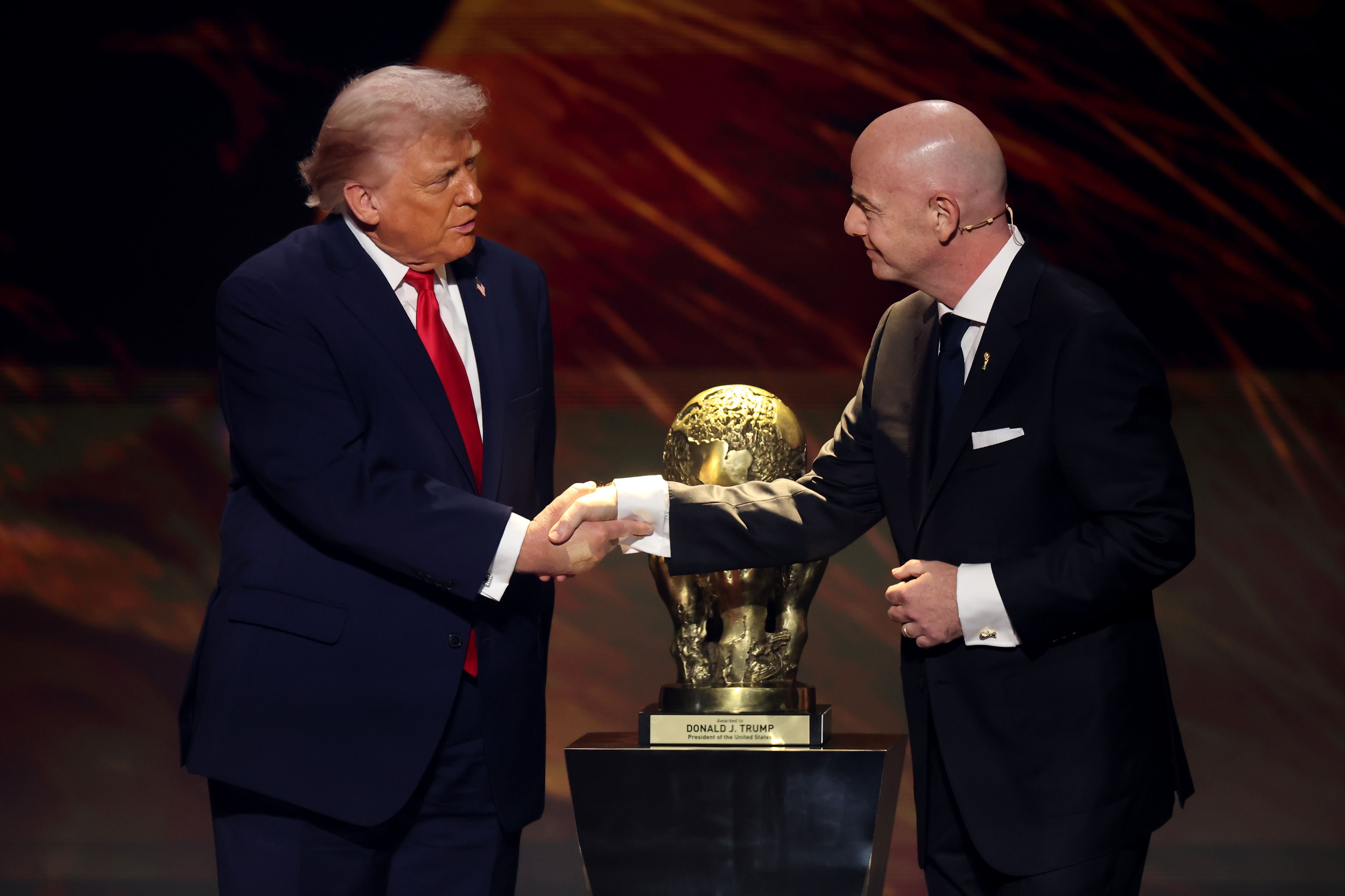 Zwei Männer im Anzug schütteln sich vor einer FIFA-Trophäe auf einer Bühne die Hand.