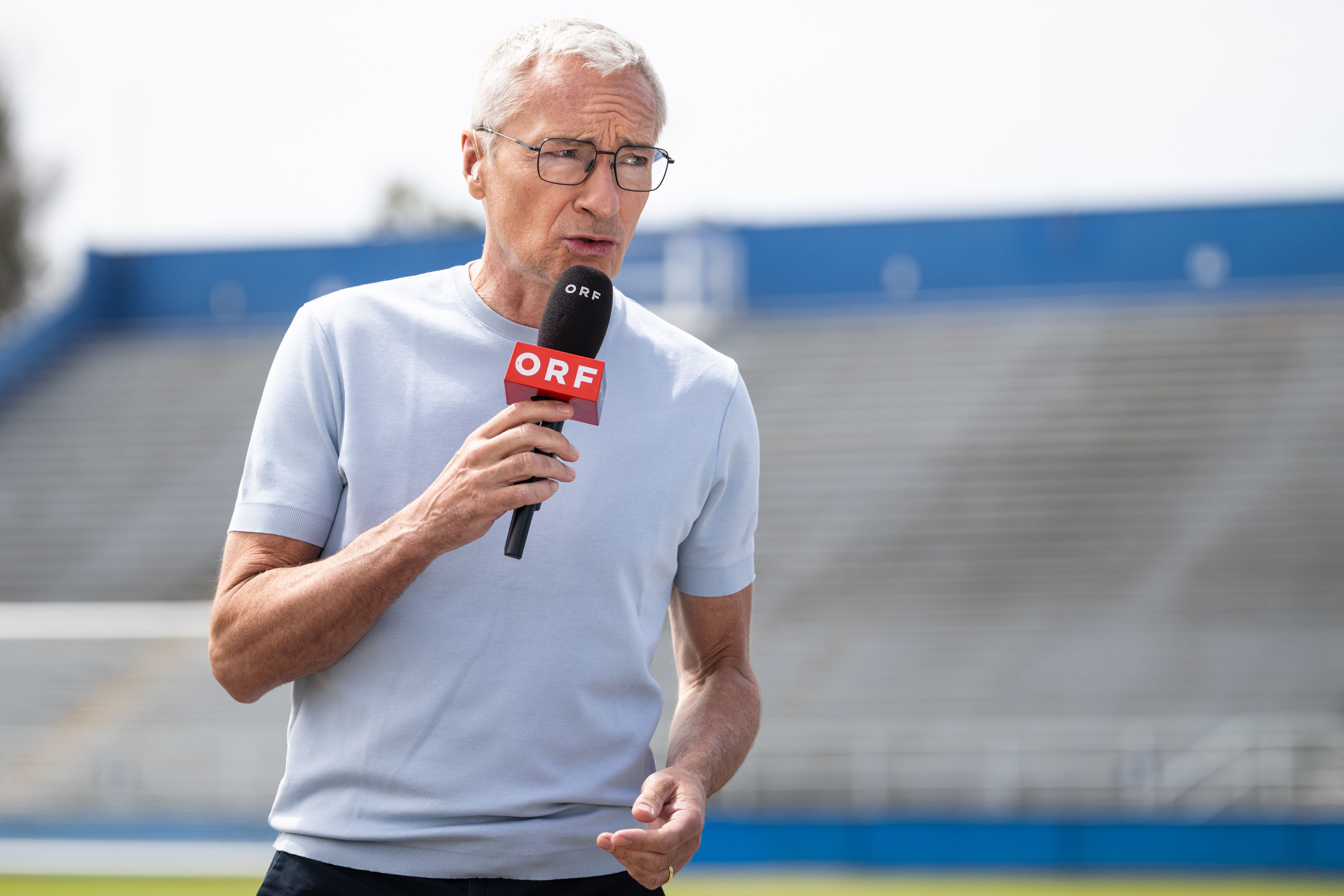 Ein Mann mit ORF-Mikrofon steht im Stadion und spricht, im Hintergrund leere Tribünen.