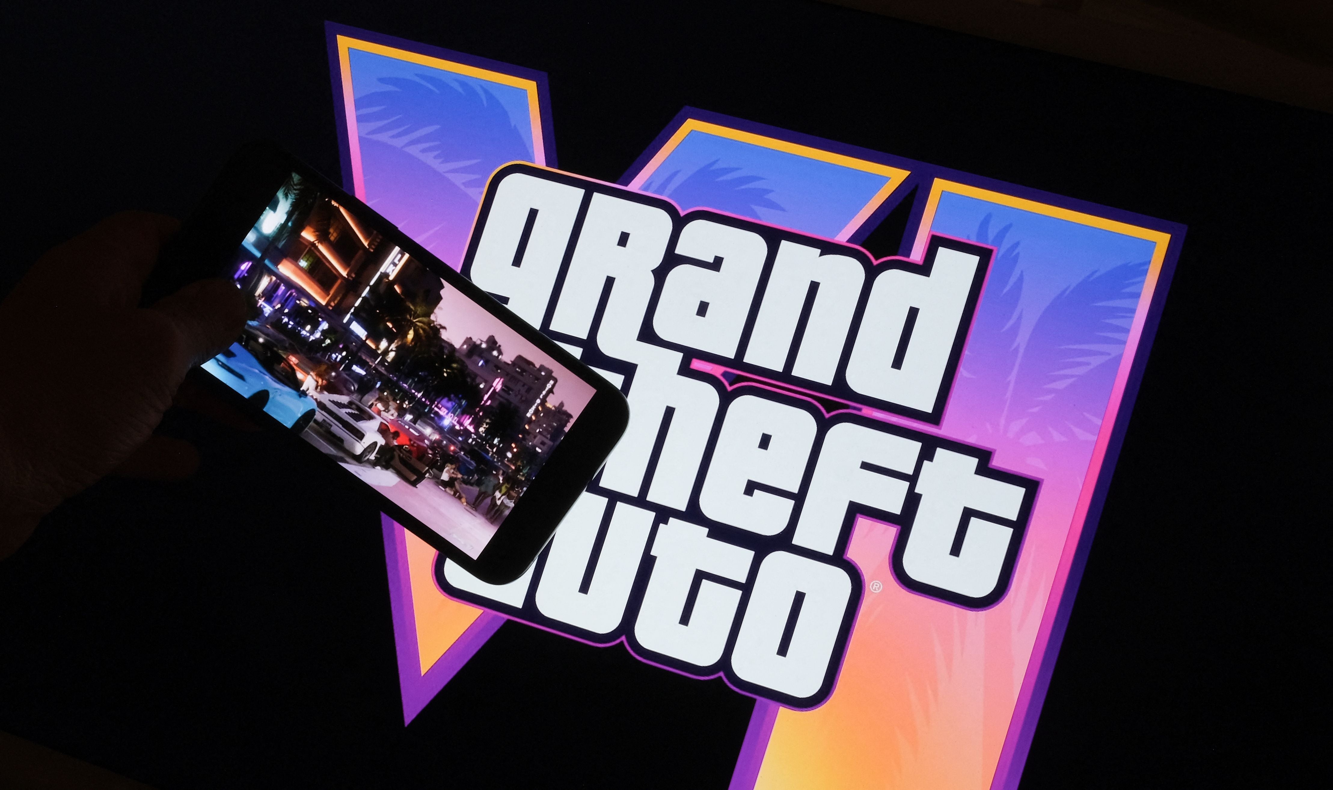 Hand hält Smartphone mit GTA VI-Trailer vor großem "Grand Theft Auto VI"-Logo auf Bildschirm.
