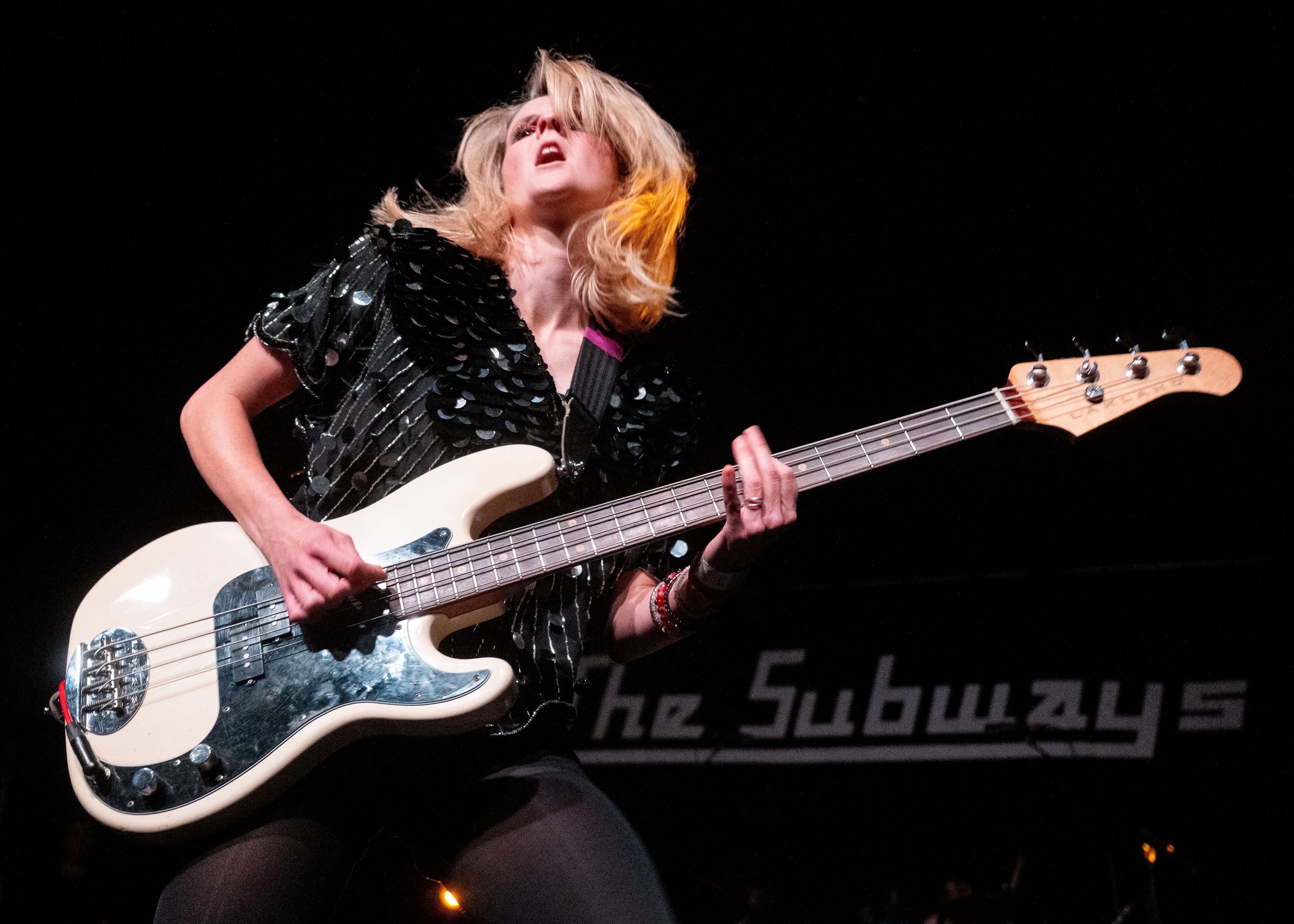 Eine Musikerin spielt Bassgitarre auf der Bühne mit einem The-Subways-Logo im Hintergrund.