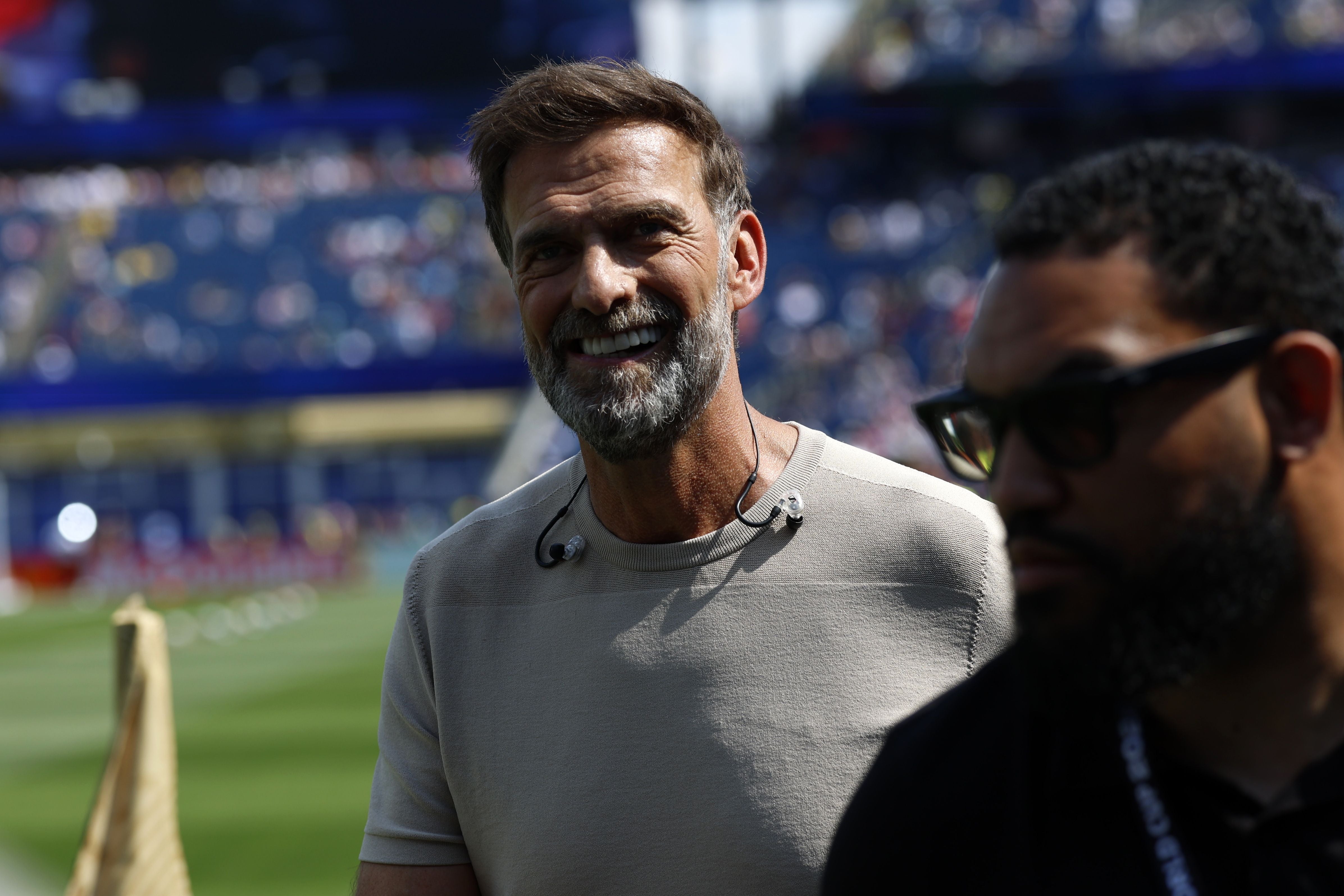 Ein lächelnder Mann steht im Stadion, im Hintergrund sind unscharfe Zuschauerränge zu sehen.
