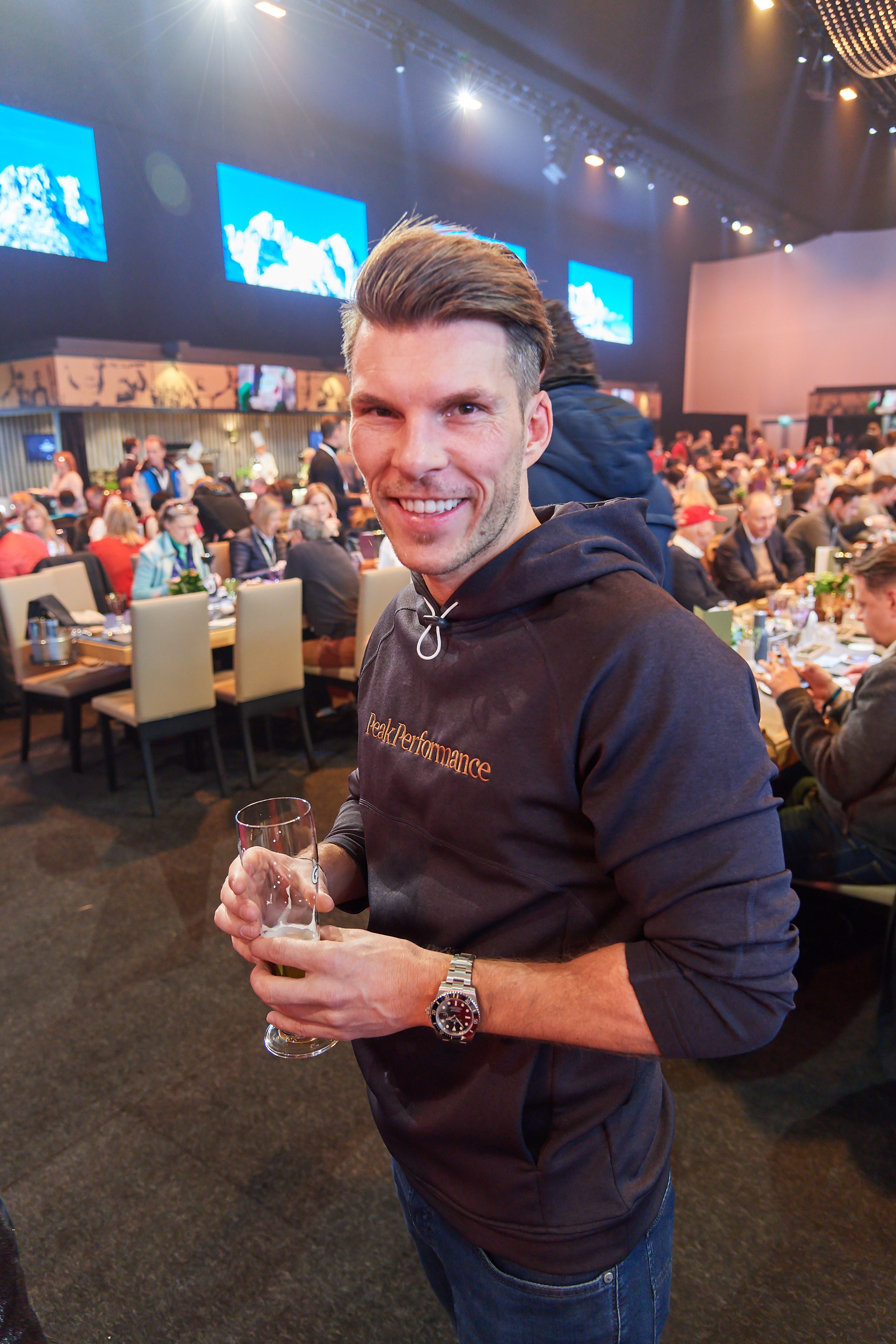 Ein Mann mit Hoodie hält ein Glas in einer vollen Veranstaltungshalle und lächelt.
