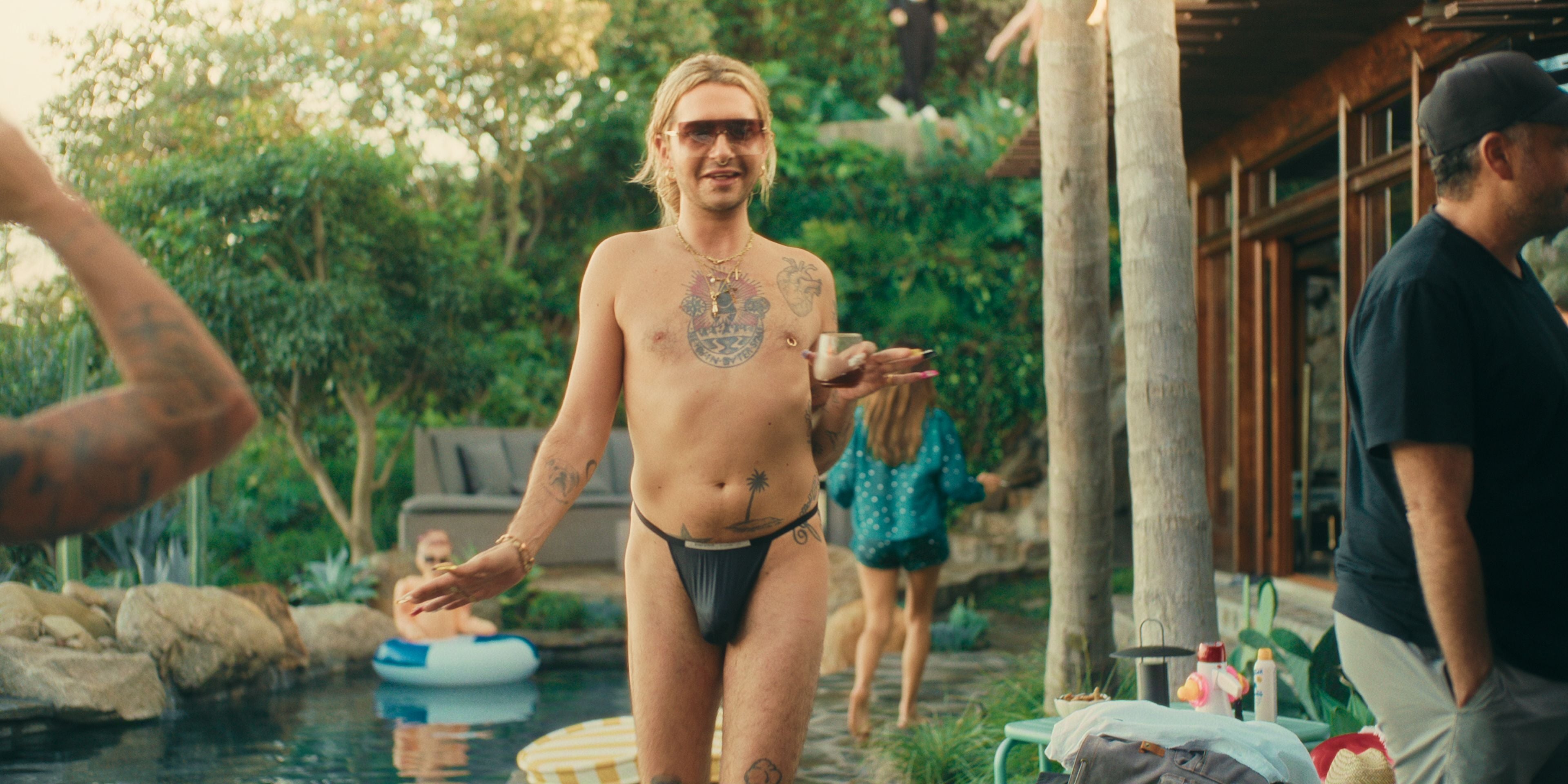 Ein Mann mit Sonnenbrille und Tattoos steht in Badebekleidung am Pool bei einer Gartenparty.