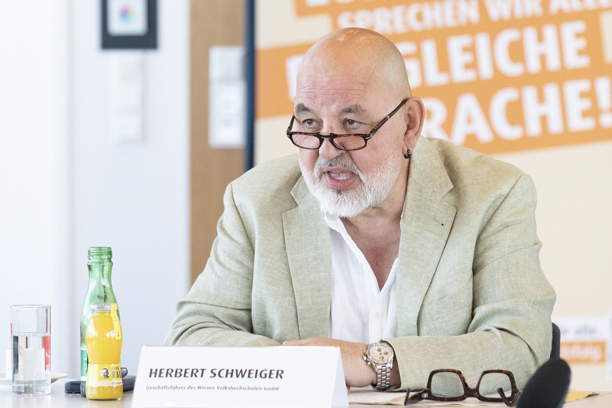Ein älterer Mann mit Brille spricht bei einer Konferenz, vor ihm ein Namensschild und Getränke.