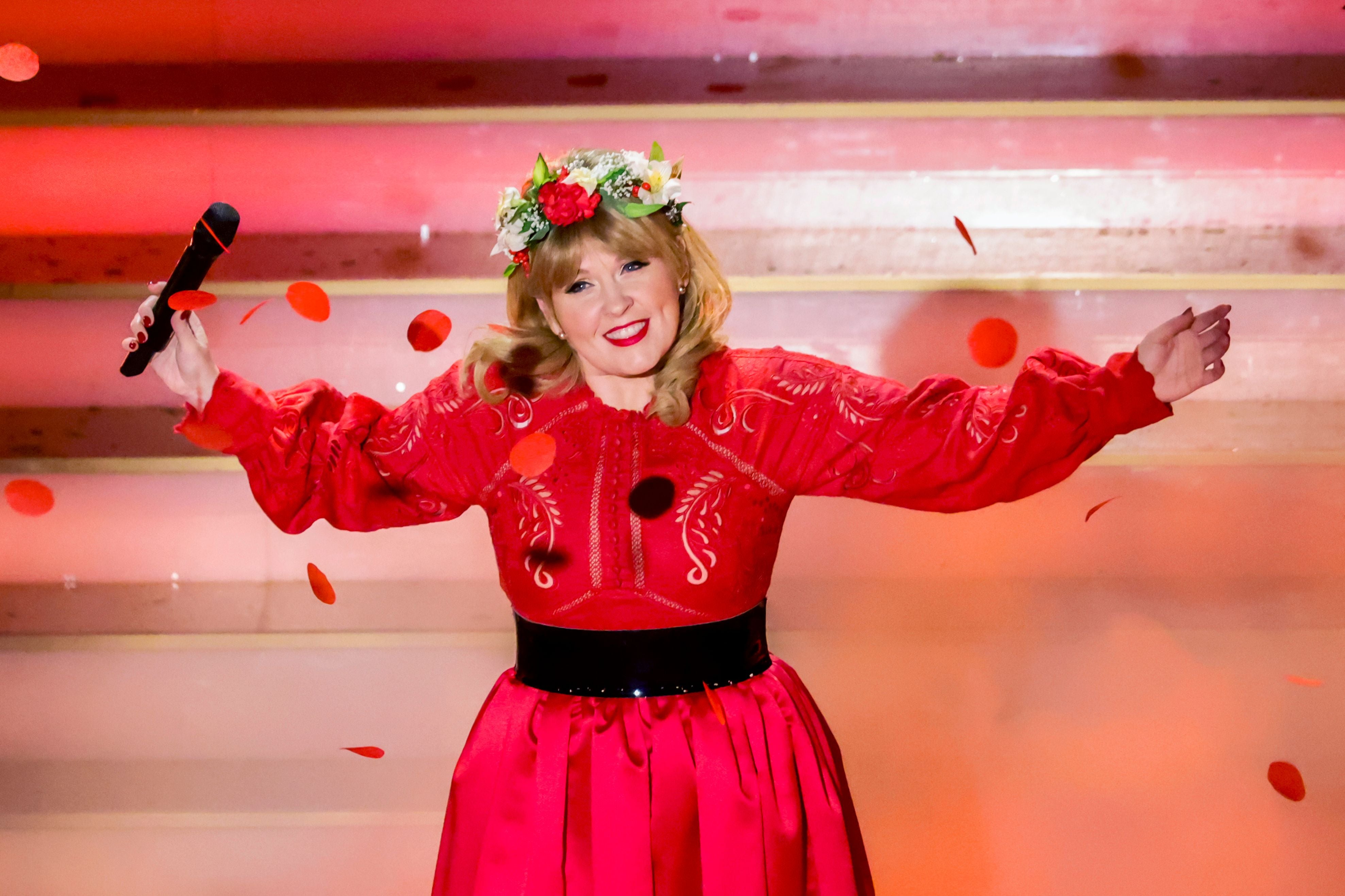 Frau in rotem Kleid mit Blumenkranz singt auf Bühne und streut rote Blütenblätter.