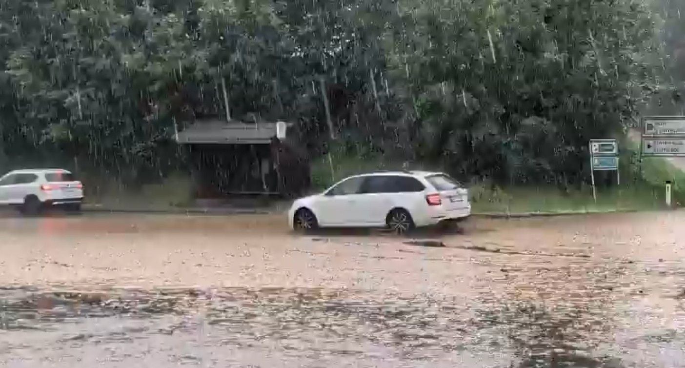 Starker Regen überflutet eine Straße, Autos fahren durch das Wasser, Bäume im Hintergrund.