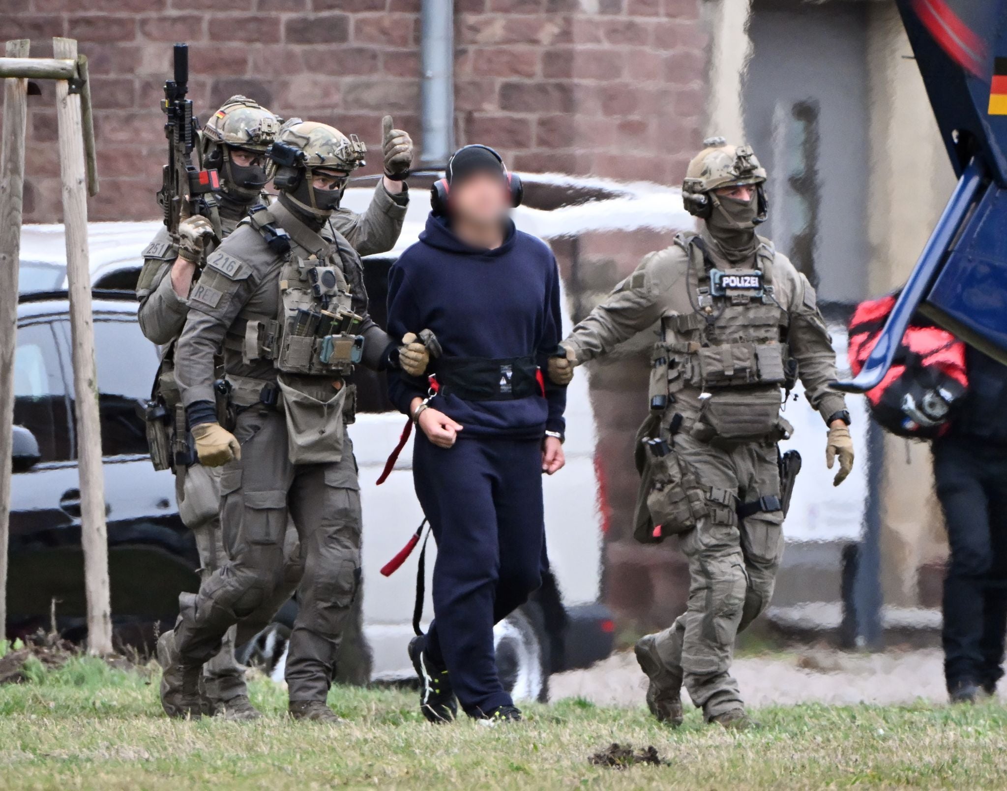Bewaffnete Polizisten führen eine gepixelte Person zu einem Hubschrauber auf einer Wiese.