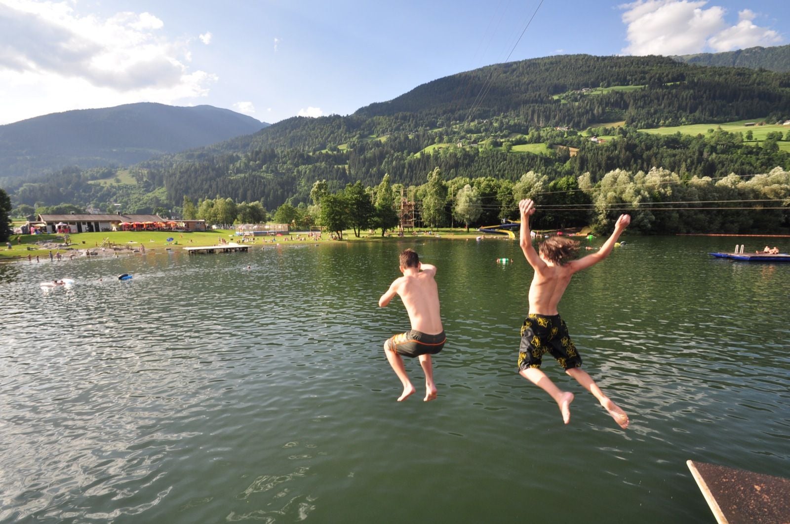 Zwei Jungen springen in den Badesee Greifenburg, im Hintergrund Berge und grüne Wiesen.
