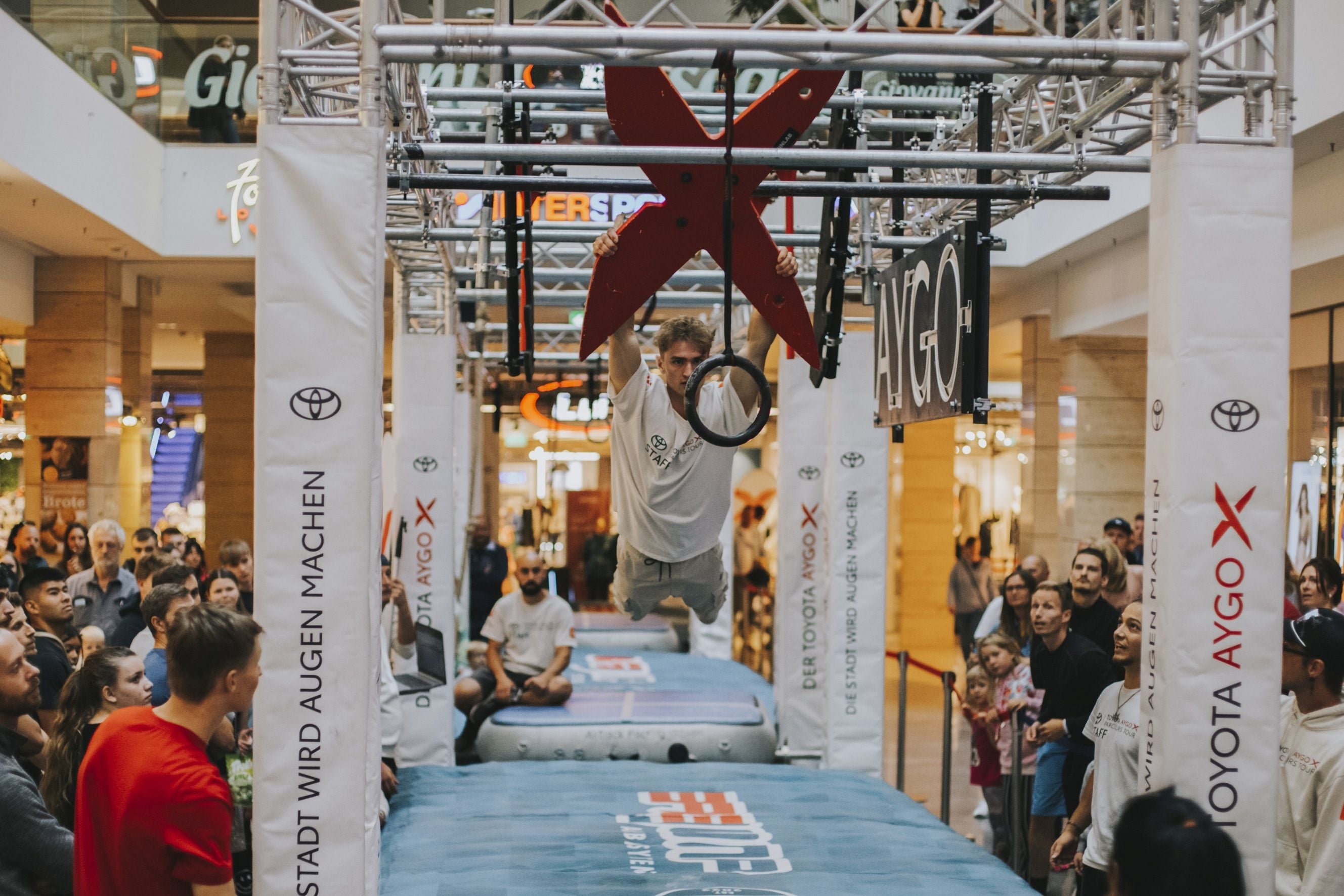 Ein junger Mann hängt an einem Ring eines Ninja-Parcours in einem Einkaufszentrum, viele Zuschauer schauen zu.