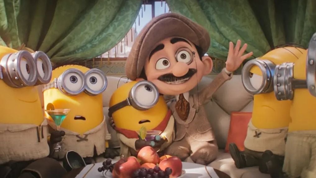 Mehrere Minions sitzen mit einem Mann in Anzug und Mütze auf einem Sofa, Obstplatte im Vordergrund.