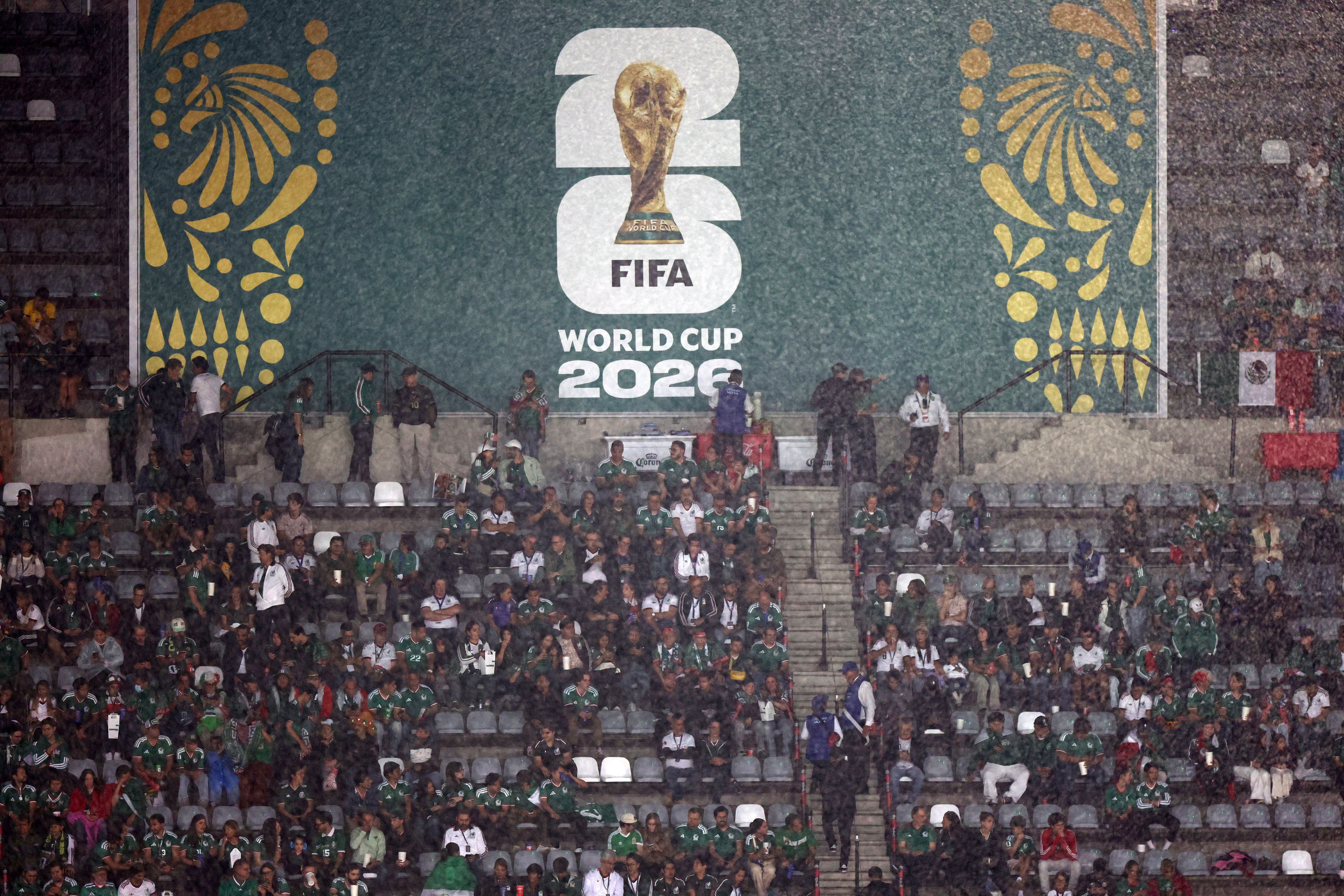 Fans im Stadion in Mexiko-Stadt warten im Regen auf ein WM-2026-Spiel, Banner im Hintergrund.