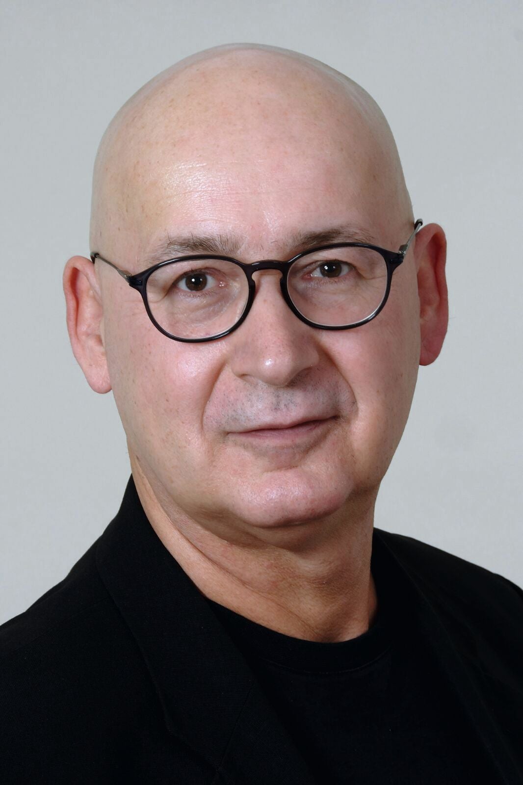 Ein glatzköpfiger Mann mit Brille und schwarzer Kleidung vor neutralem Hintergrund.