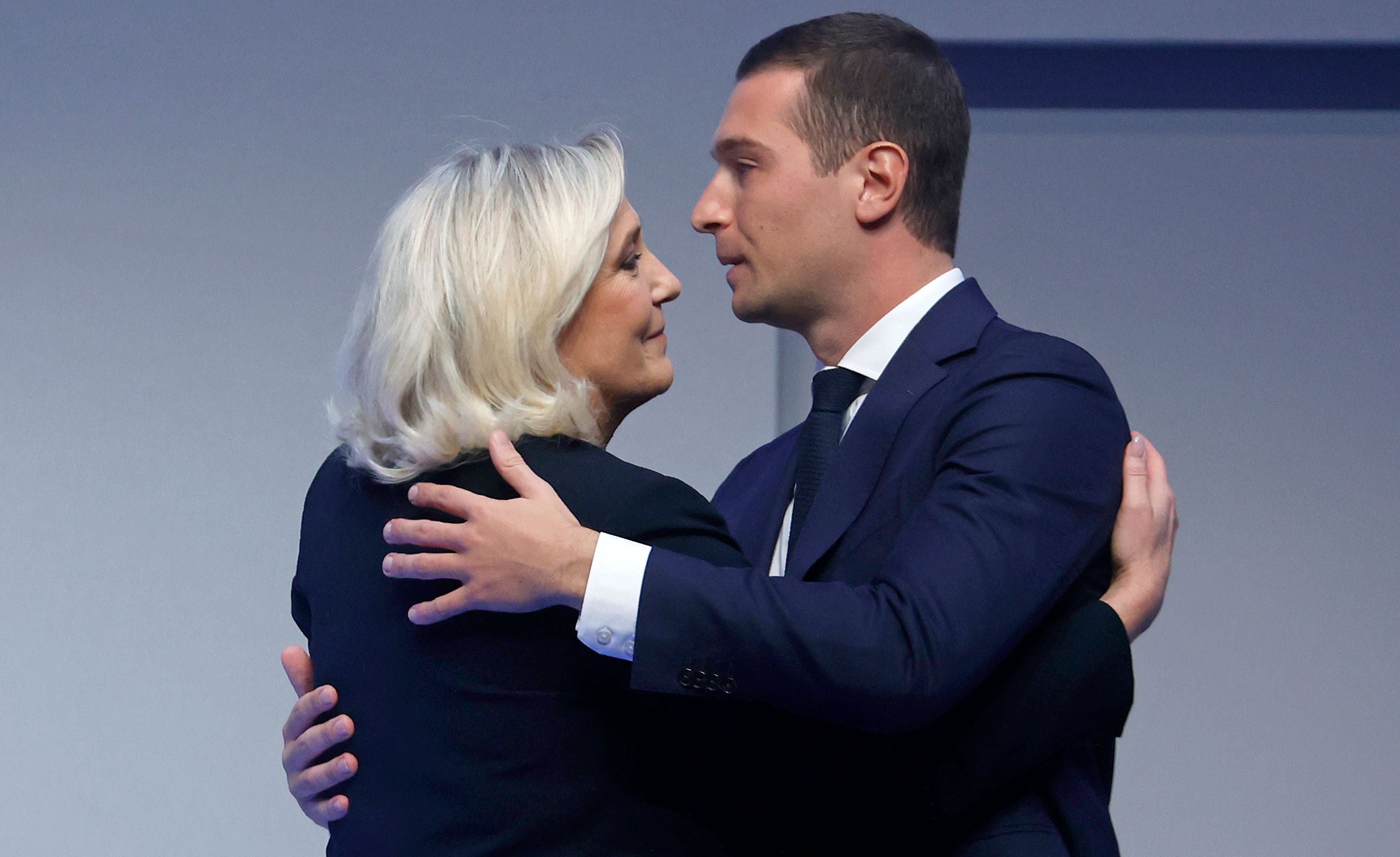 Marine Le Pen und Jordan Bardella umarmen sich auf einer Bühne vor neutralem Hintergrund.