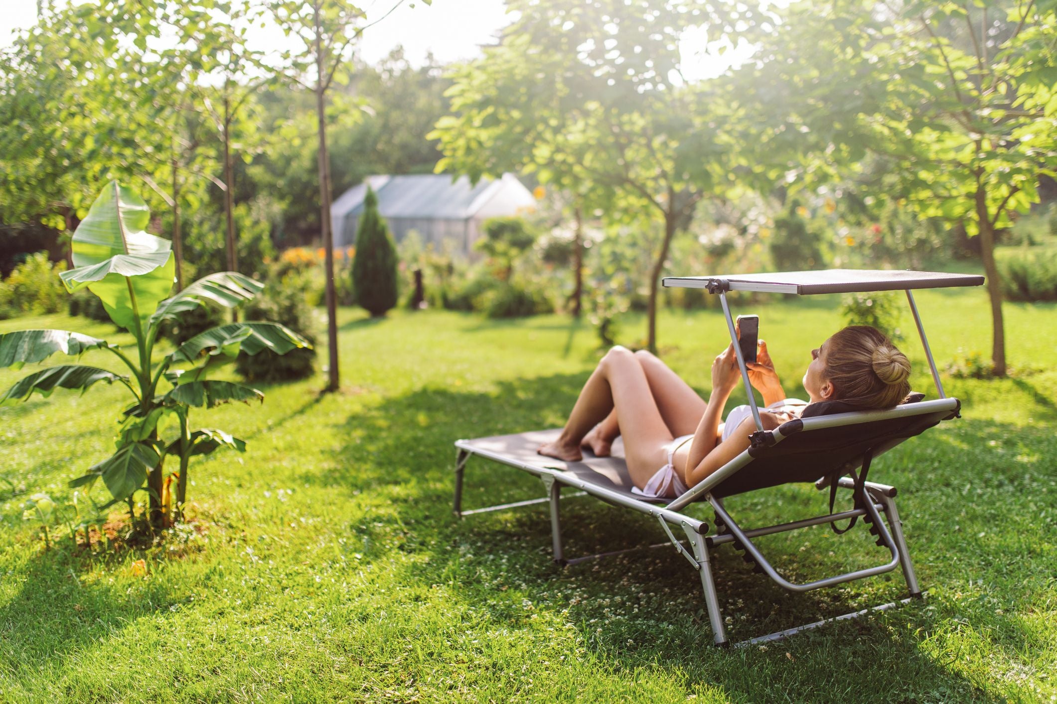 Frau liegt im Bikini auf einer Sonnenliege im Garten und benutzt ihr Handy umgeben von Grün.