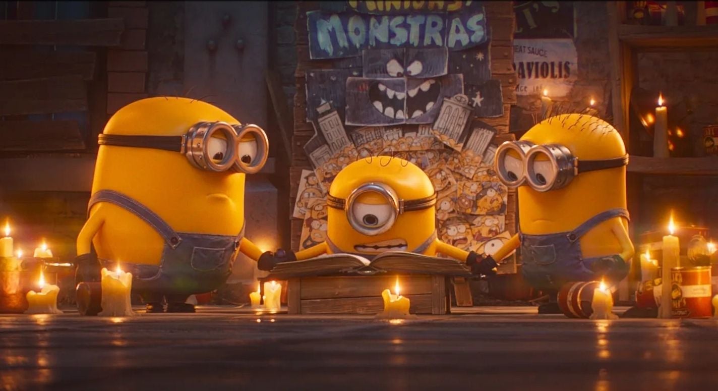 Drei Minions sitzen im Kreis, umgeben von Kerzen, und lesen gemeinsam in einem Buch.