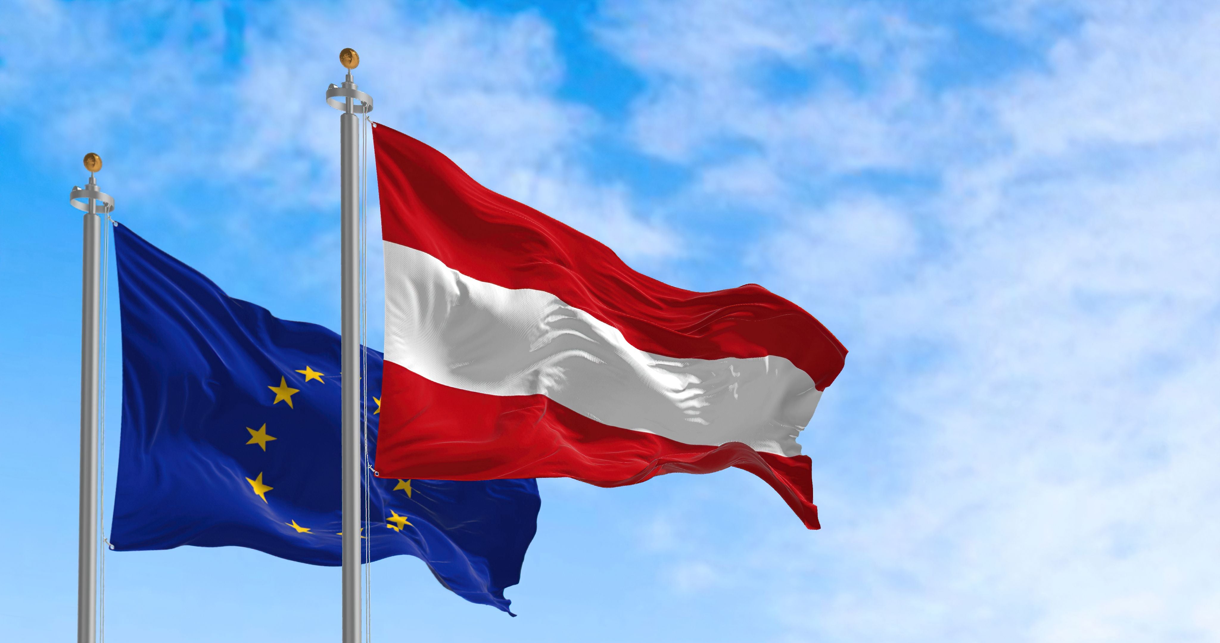 Die Flaggen von Österreich und der EU wehen im Wind vor blauem Himmel.