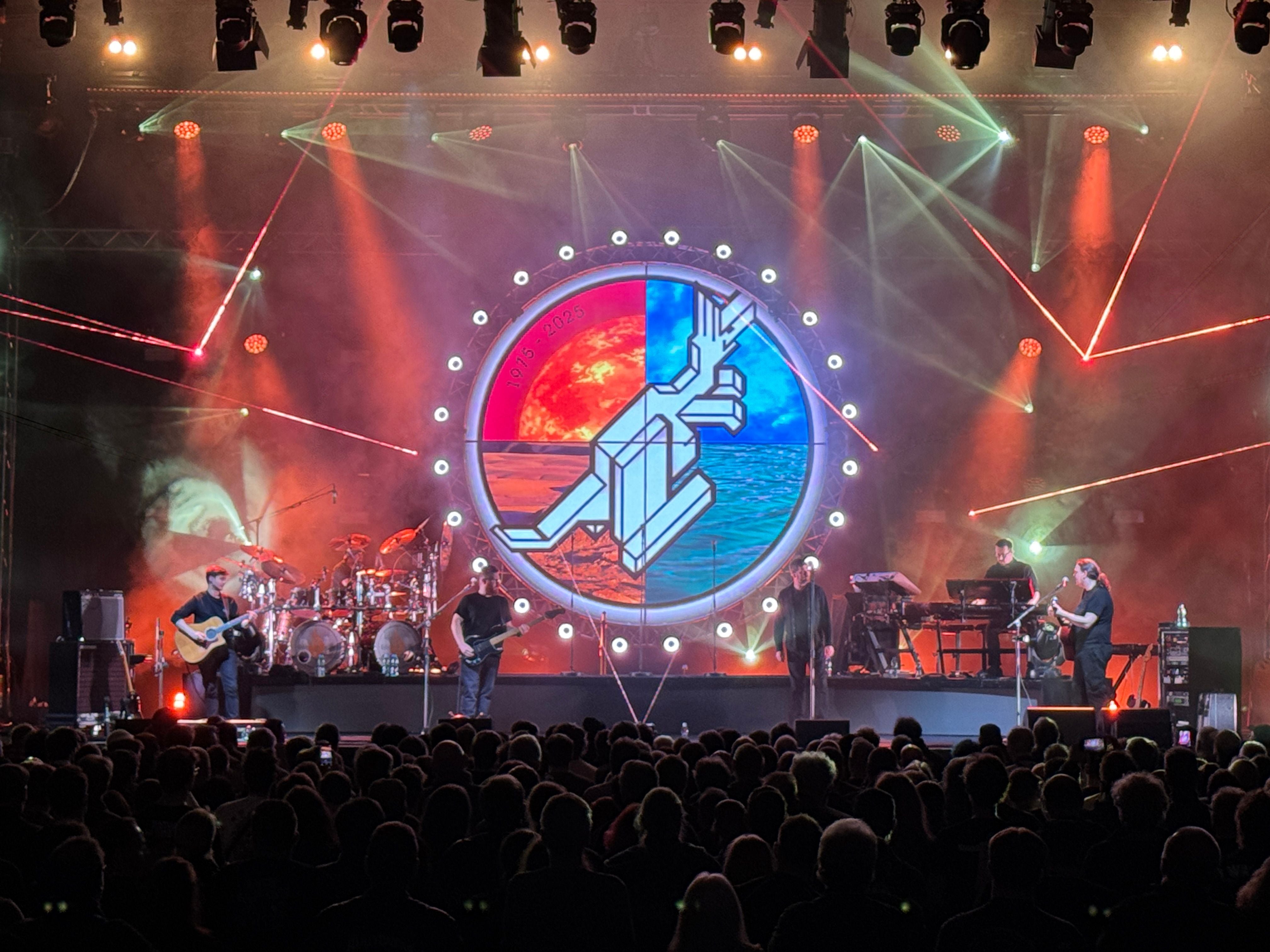 Eine Band spielt live auf einer Bühne mit bunten Lichtern und großem runden Leinwand-Hintergrund.