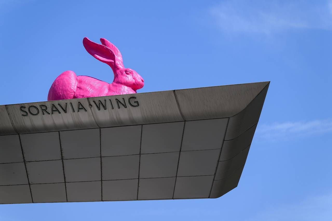 Große pinke Hasenskulptur sitzt auf dem Dach des Soravia Wing vor blauem Himmel.
